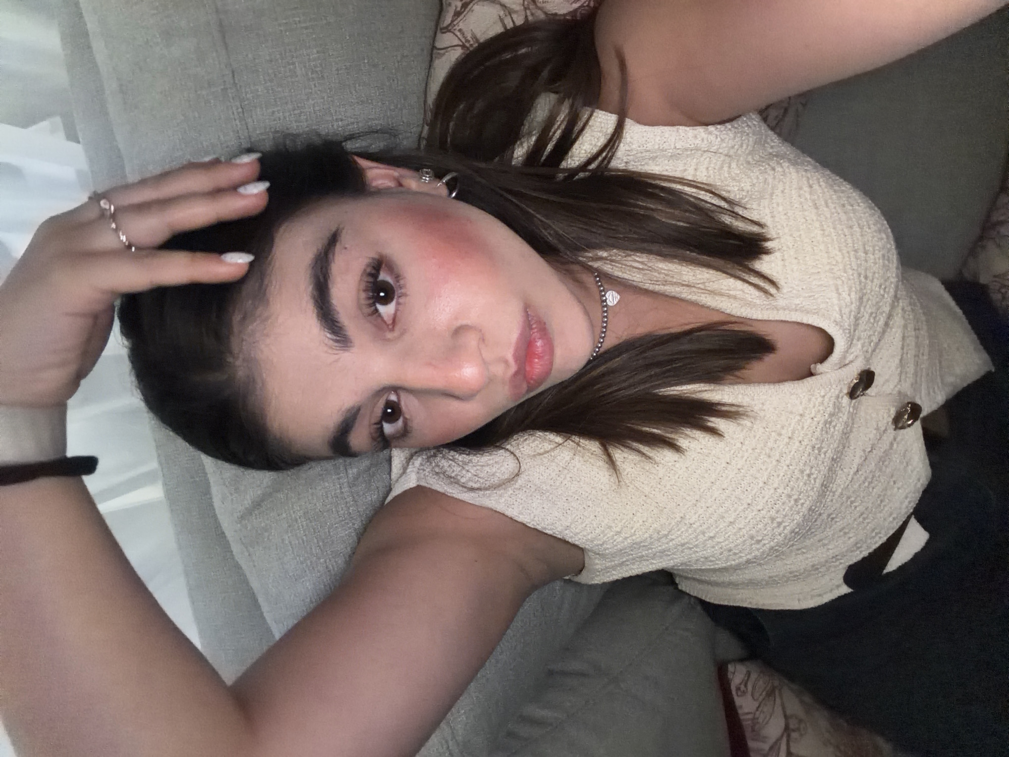 A young woman lies on a gray cushioned surface, possibly a sofa, indoors. She is wearing a light-colored, textured top with button details and dark pants. Her hair is brown, and she wears a necklace and earrings. Her right hand is raised towards her head. The background is blurred, and there is no immediate indication that the location is Barrio de Nuevo León, Mexico.
