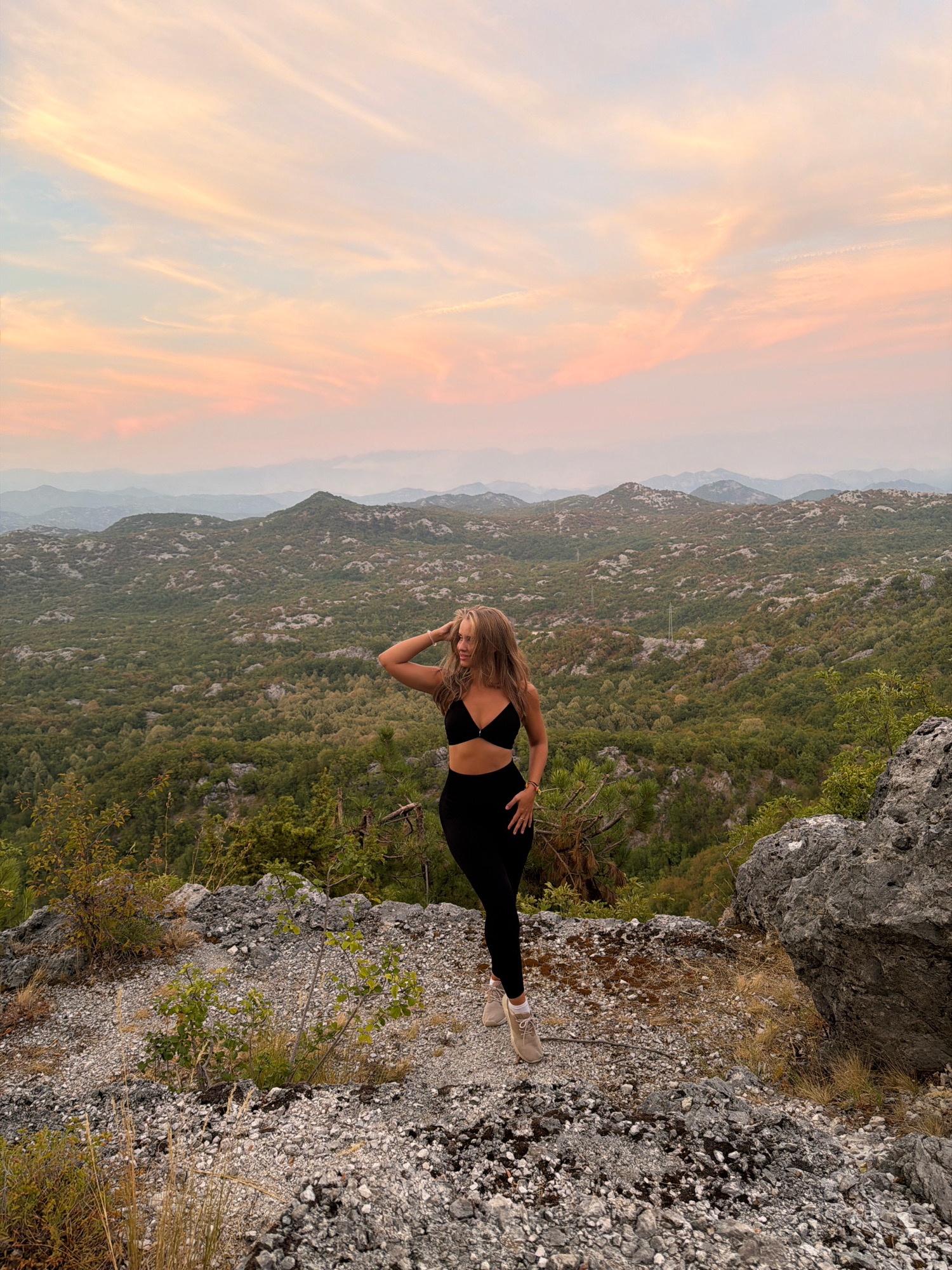 A young woman stands on a rocky outcrop overlooking a lush, green mountainous landscape at dusk. She is wearing a black sports bra and black leggings. Her right hand is raised to her head, and her left hand is on her hip. The sky is a soft blend of pink, orange, and blue, indicating either sunrise or sunset. The mountains in the background are covered in dense vegetation, with rolling hills stretching into the distance. The foreground is rocky and uneven, with scattered dry grass and small bushes. The overall impression is one of serene natural beauty. This scene is located in the mountainous terrain of Boguti, Montenegro.