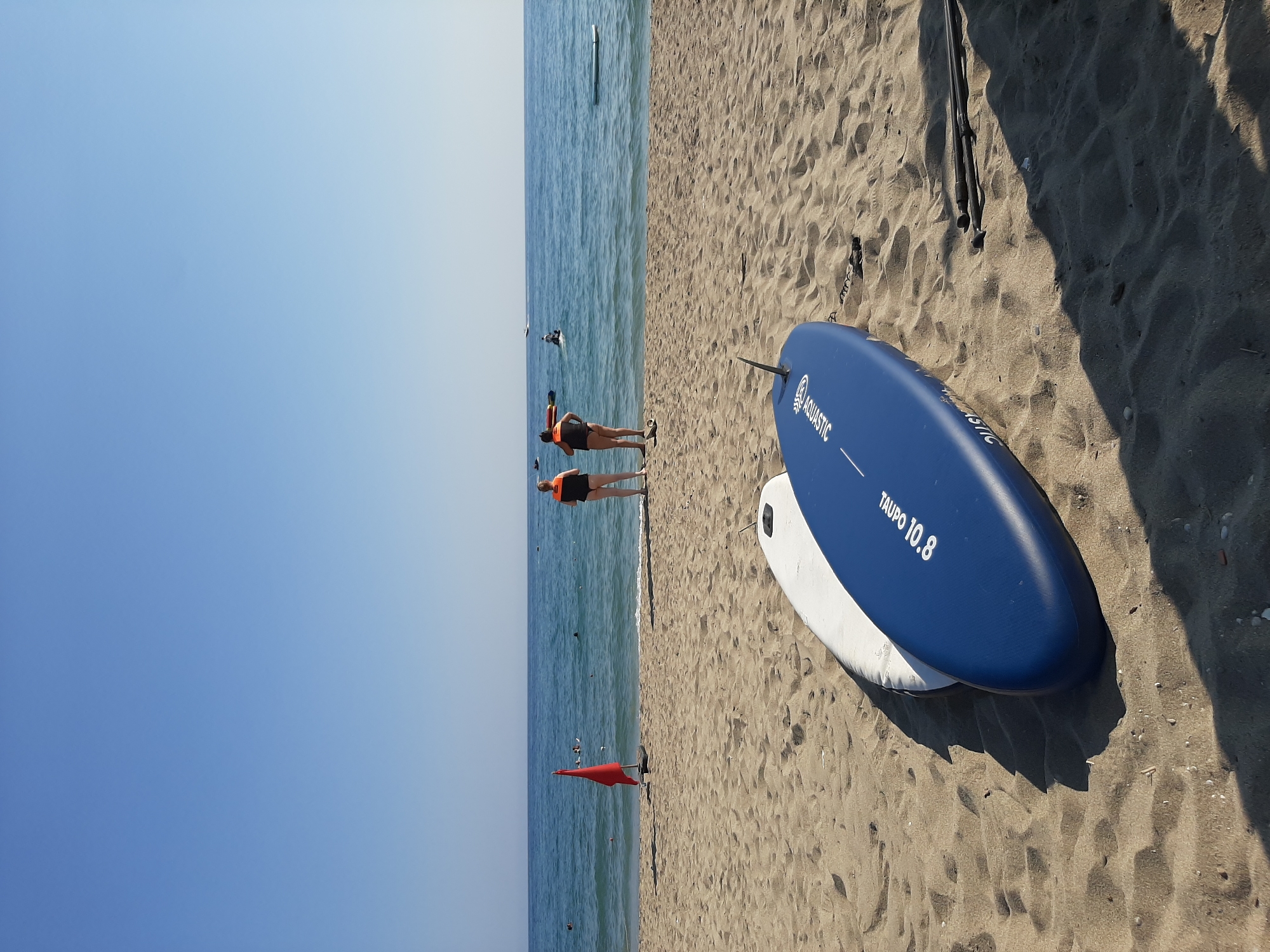 The image depicts a sandy beach scene on a clear, sunny day, likely in Alanya, Turkey. Two stand-up paddleboards, one white and one blue, are positioned on the sand in the foreground, with the blue board closer to the viewer and bearing the text "TAUPO 10.8" and "AQUASTIC". Two people, a male and a female, both wearing life vests, are standing on the sand near the water's edge, facing the sea. The sea is calm with a few other individuals visible in the water, some on paddleboards or similar floating devices. A red flag is planted in the sand to the left of the two people. The background is a clear blue sky and the horizon where the sea meets the sky. The overall environment suggests a recreational area at the seaside.