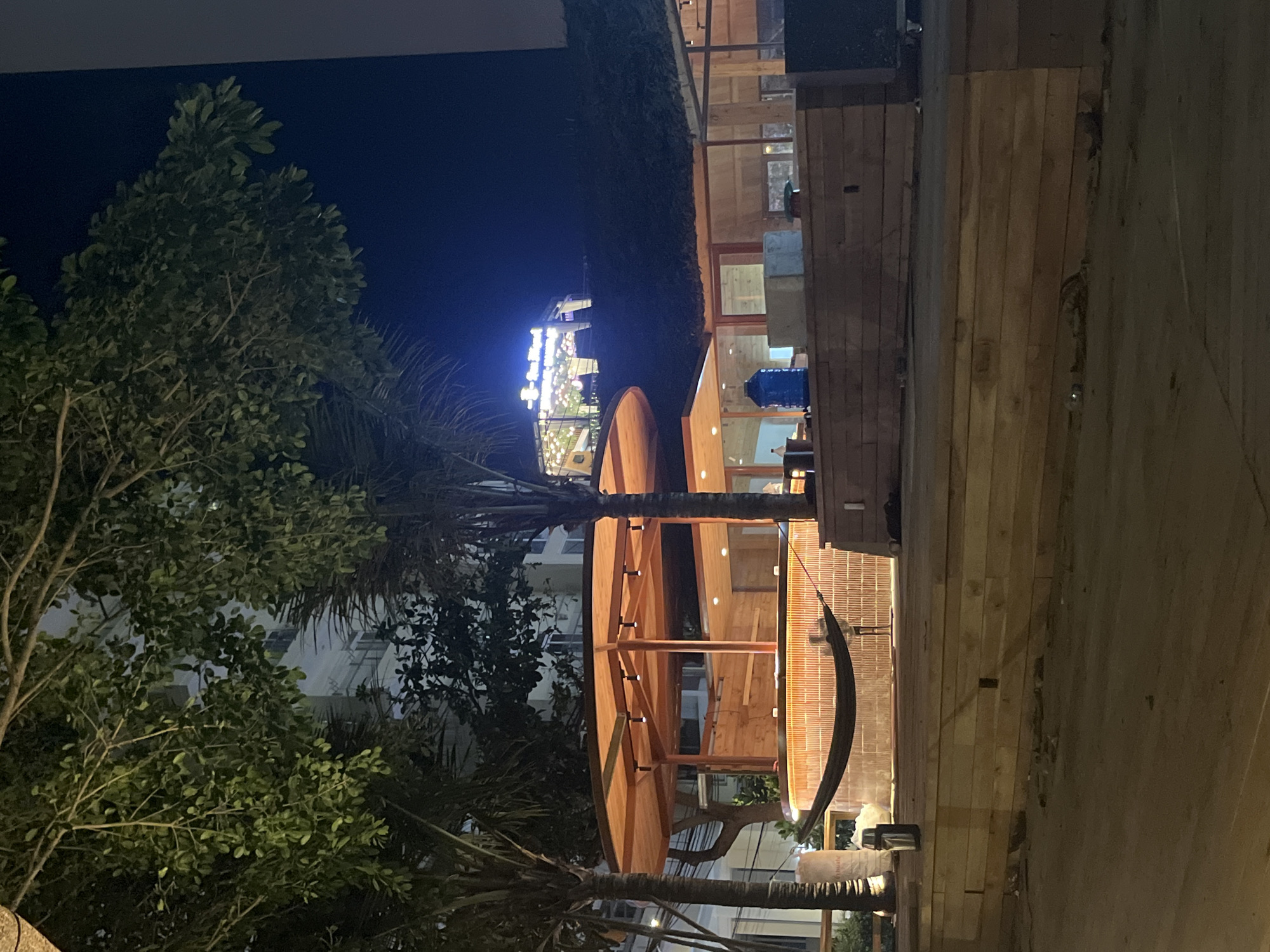 Captured from an angle, the image reveals an outdoor setting during nighttime in the city of Bac Nha Trang Ward, Vietnam. On the right side, there is a wooden structure, possibly a platform or deck. To the left, there are lush green trees and palm trees. In the distance, there are lights and buildings, indicating the presence of urban development. The overall ambiance suggests a tropical setting.