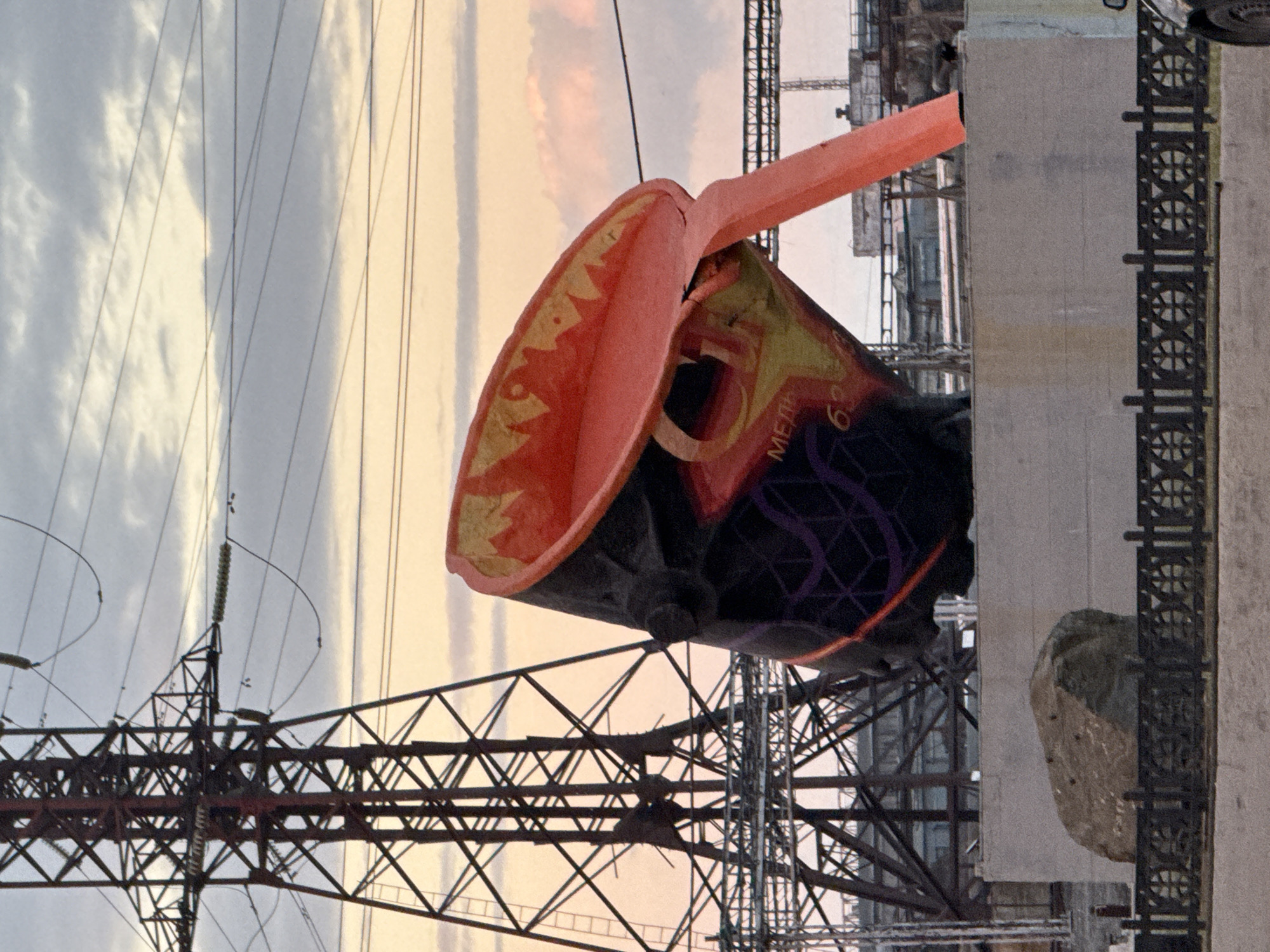 The image depicts a large, industrial-style sculpture, resembling a ladle or crucible, in Norilsk, Russia. The sculpture is predominantly black with a vibrant red interior and yellow accents around the rim, featuring a pouring spout. Some Cyrillic lettering, "МЕДЬ" (MED', meaning COPPER) and "63", is visible on the sculpture's surface. In the background, a massive electrical transmission tower with multiple crossbars and insulators dominates the left side of the frame. A series of overhead power lines crisscross the sky. The setting appears to be outdoors, with a cloudy sky exhibiting hues of orange and pink, suggesting either sunrise or sunset. The sculpture is situated on a raised concrete platform, and in the foreground, a large, rough-hewn rock rests on the ground. A decorative black metal fence runs along the edge of the foreground area. The overall scene conveys an industrial aesthetic, likely in an urban or industrial zone of Norilsk, given the presence of the power infrastructure and the sculpture's theme. There are no people visible in the image.