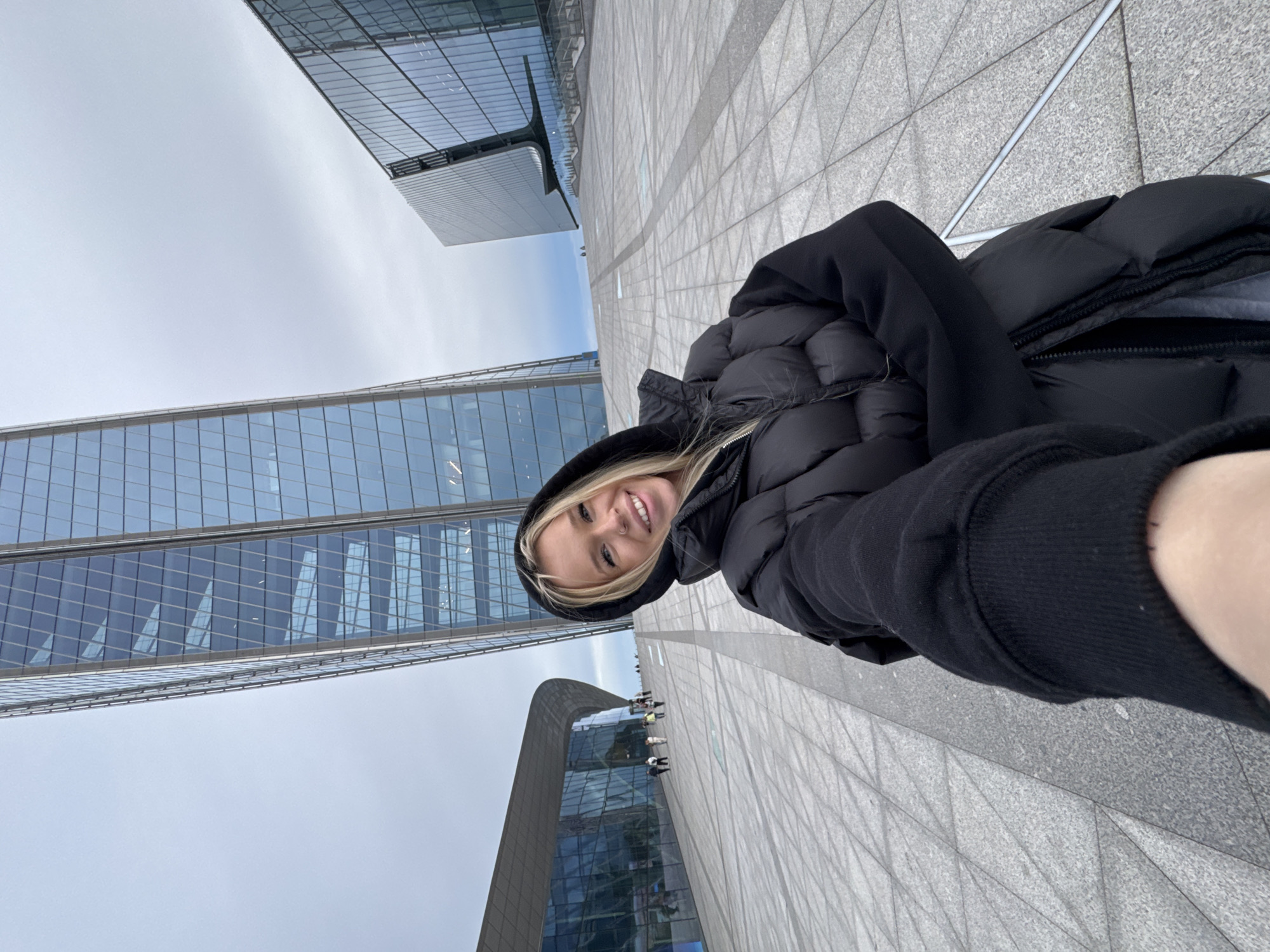 A young blonde woman with a nose ring is taking a selfie in front of modern glass buildings under a cloudy sky. She is wearing a black hooded puffer jacket. The woman is smiling and looking at the camera. The background features a tall, leaning skyscraper with a glass facade and another, more angular glass building. The ground is paved with large grey tiles. Several other people are visible in the distance, walking on a wide, sloped walkway. The scene appears to be a public plaza or architectural complex in a city. This location is the Lakhta Center in Saint Petersburg, Russia.