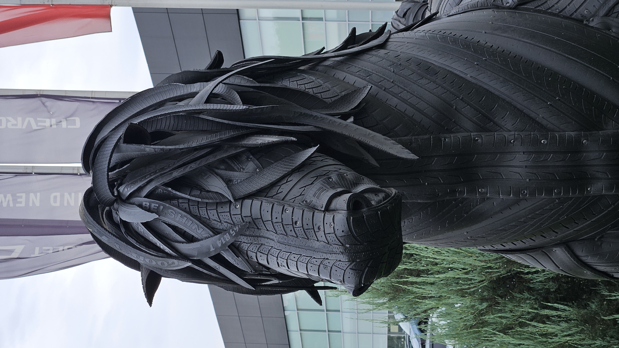 An outdoor, medium shot captures a black sculpture of a horse's head and upper torso, crafted entirely from recycled tires. The horse's mane is depicted as flowing, layered strips of tire treads, with some of the tire branding, such as "GOOD YEAR" and "BELSHINA," visible. The horse's face is intricately formed from tire sections, showing the texture of the treads and the embedded metal studs. To the left of the sculpture, a dense, green evergreen bush is partially visible. In the background, a modern, dark-paneled building with large, reflective windows stands under a cloudy sky. Several large, purple banners with white lettering hang from poles to the left of the building. One banner clearly displays the word "CHEVROLET" in large, capitalized letters. The location is the Chevrolet dealership in Almaty, Kazakhstan.