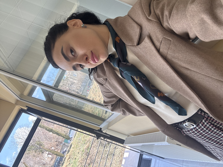 A female individual is photographed in a selfie-style image indoors, positioned in front of a large window or glass enclosure. She has dark hair pulled back, prominent eyebrows, and wears pink lipstick, exhibiting slightly pouted lips. She is dressed in a light-colored turtleneck, a dark green patterned scarf, a light brown blazer, and a dark and white patterned skirt secured by a belt with a white double-ring buckle. A white radiator is partially visible on the interior wall in the lower left.
Beyond the window, an outdoor residential environment is visible under a clear blue sky with scattered clouds. The terrain features a gently sloping hillside covered in dry, light-colored vegetation and numerous deciduous trees with bare branches, indicative of a colder season. A road or parking area is visible with multiple parked vehicles, including at least two dark-colored cars and one lighter vehicle. In the background, several multi-story residential buildings, consistent with Soviet-era architecture, are observed. The scene depicts a residential district within Tbilisi, Georgia, characterized by its hilly landscape and urban development.