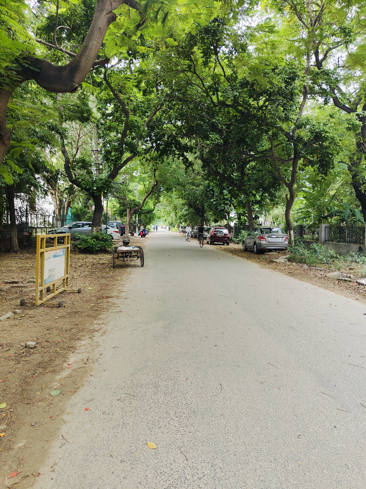 This image depicts a street scene in Gurgaon, India, characterized by a tree-lined road with dense foliage casting shadows. On the left side of the road, a yellow barrier with wheels and a white rectangular sign is positioned on a dirt and leaf-covered area. A few parked cars are visible further down the road. In the foreground, a small, two-wheeled cart with a flatbed is present. As the road recedes into the distance, more trees and vegetation line both sides, with a few individuals on bicycles and a motorcycle visible. The road surface is asphalt. The overall environment suggests a moderately busy urban street with significant greenery.
