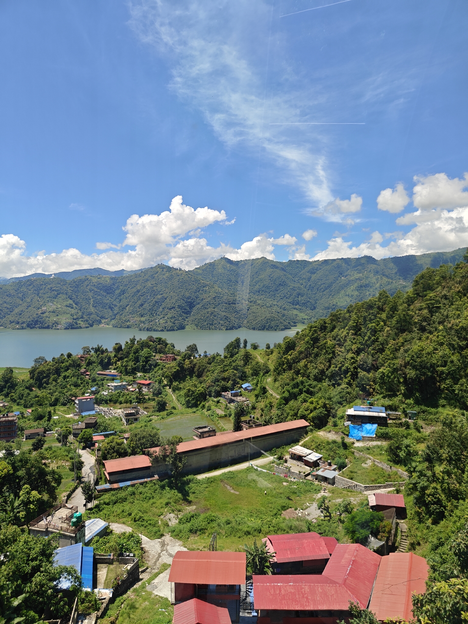 This is a high-angle, medium shot of a landscape in Pokhara, Nepal, featuring a large body of water, rolling green hills, and a cluster of buildings. The sky is a bright blue with scattered white clouds and two thin white contrails from aircraft. The foreground is dominated by lush green vegetation and several buildings with red or blue roofs. The buildings appear to be houses or small commercial structures. Some structures have visible solar panels. A paved road winds through the scene, connecting some of the buildings.
In the middle ground, a significant body of water, likely a lake, stretches across the image, reflecting the blue of the sky. Beyond the lake, a series of densely forested hills rise, forming a backdrop. The hills are covered in a vibrant green, indicating a temperate climate. The overall impression is one of a serene, natural setting interspersed with human habitation. There are no people clearly visible in the image.
