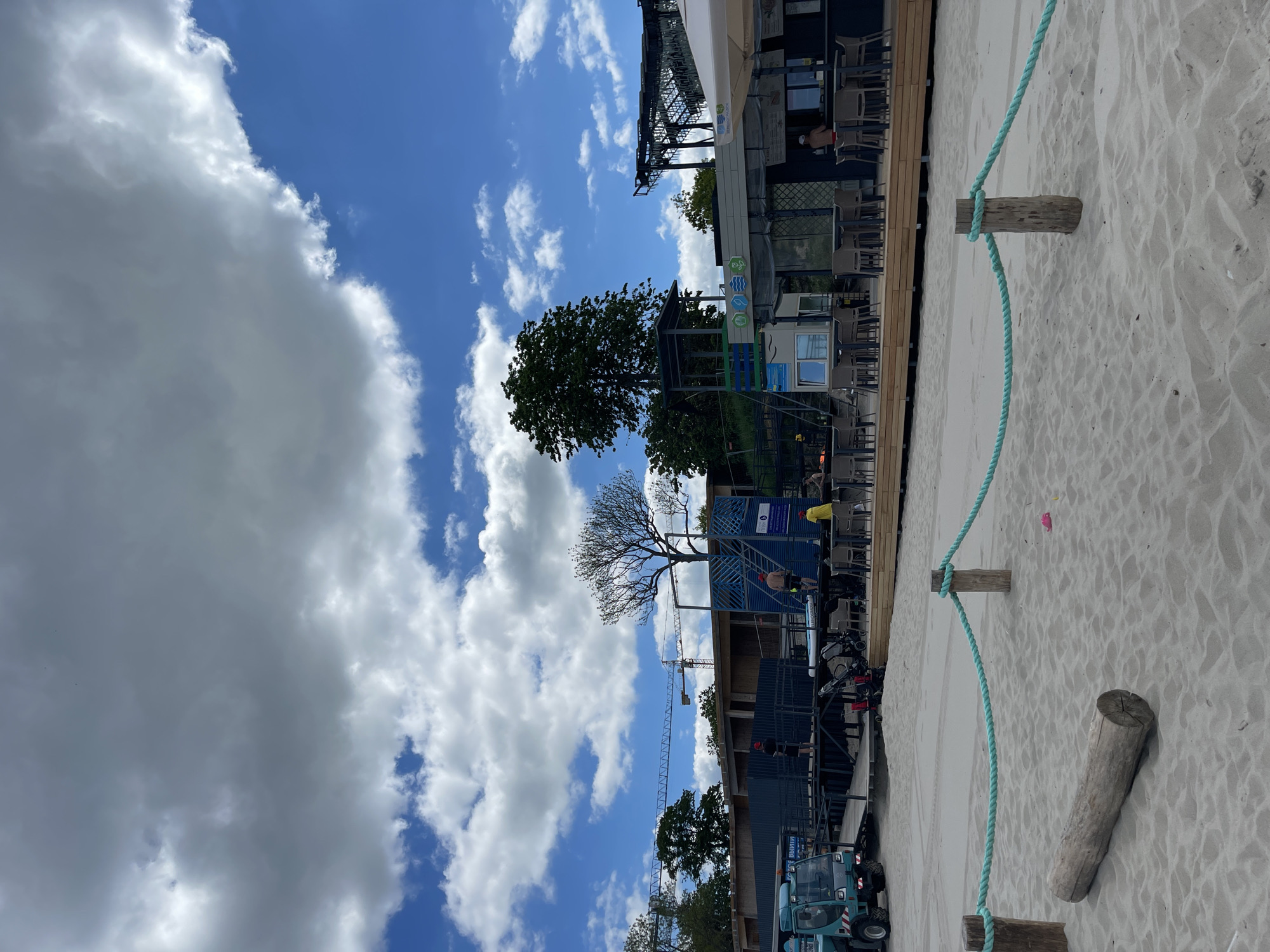 The image displays a lively beachside area in Zelenogradsk, Russia, under a bright, partly cloudy sky. The scene is oriented as if rotated 90 degrees clockwise, placing the sky at the top, the beach on the right, and buildings/structures on the left.
On the right side, a sandy beach is visible, separated from the built-up area by a light blue rope. This rope is strung between three simple wooden posts embedded in the sand, while a fourth wooden log lies horizontally on the beach. A small, pink object, possibly a child's toy, rests on the sand near the rope.
To the left, a series of structures, likely beach cafes or commercial establishments, line the sand. These buildings feature outdoor seating with tables and chairs, some protected by a white awning. Several people are present; one individual in a red top and hat is seated at a table, and another in a yellow top is visible further along. A blue barrier or fence with some illegible symbols (including waves) marks part of these structures.
In the mid-ground and background, a variety of trees are present, ranging from full, leafy green trees to one striking bare-branched tree. Evidence of construction is apparent with a large crane extending into the sky, and a blue construction vehicle, possibly a telehandler, is parked near the buildings. The vehicle has partially visible Cyrillic text that appears to say "ВЫШЛИФУ" (Vyshlifu).
The sky dominates the upper portion of the image, showing large, fluffy white and grey cumulus clouds interspersed with clear patches of vivid blue. The weather appears fair, suggesting a pleasant daytime setting for beach visitors and ongoing activities.