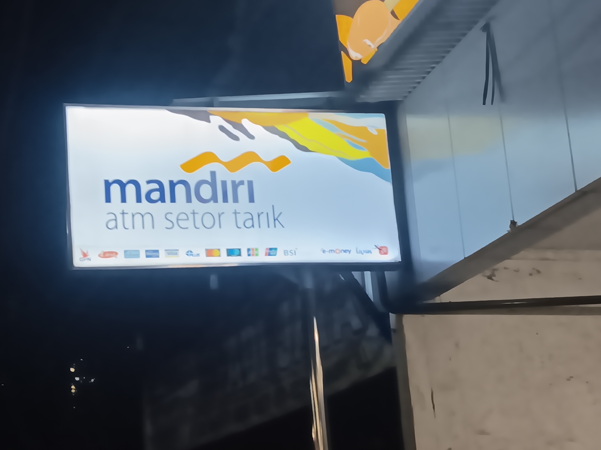 The image shows a brightly lit, rectangular sign at night, likely in Batursari, Indonesia, displaying "mandiri atm setor tarik" which translates to "Mandiri ATM deposit withdrawal." The sign features the Mandiri logo, a stylized orange wave, and a colorful abstract graphic in yellow, blue, and gray. Below the main text are logos of various payment networks including GPN, Prima, ATM Bersama, Visa, Mastercard, and others. To the right of the payment logos, "BSI" and "e-money" are visible. The sign is mounted on a pole, and the background is dark, suggesting it's outdoors at night. The environment appears to be urban, with hints of a building and possibly some street lights in the distance.