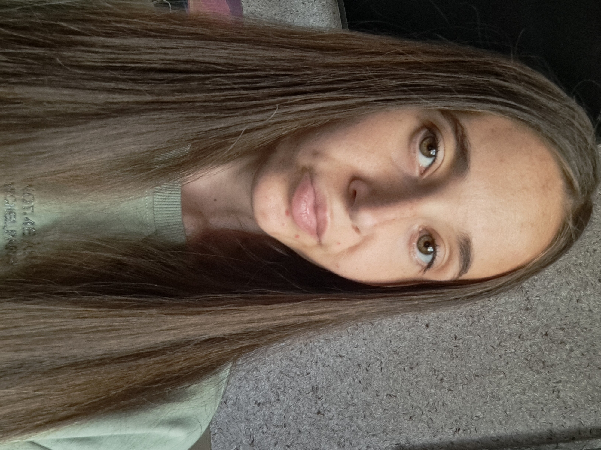 This is a head-and-shoulders, frontal, selfie-style photograph of a young woman with long, straight, light brown hair. She is looking directly at the camera with a neutral expression. Her skin is fair with some freckles and blemishes. She is wearing a light green crewneck sweatshirt. The background appears to be a textured gray wall. The image appears to have been taken indoors, likely in a residential setting. There is no identifiable information within the image to suggest a specific location in Vypasne, Ukraine.
