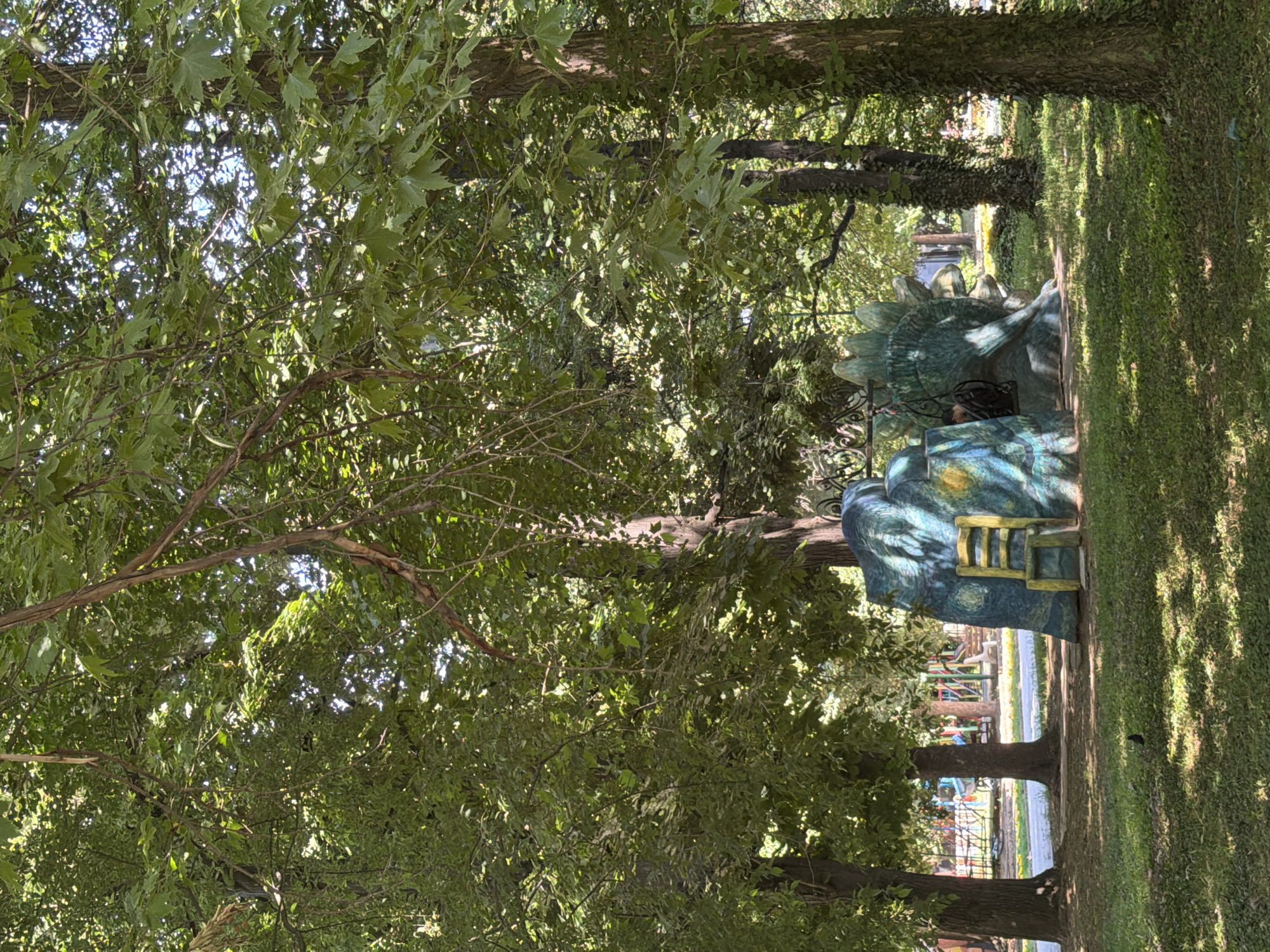 The image depicts an outdoor scene within a park. In the foreground, a grassy area is partially shaded by the dense foliage of trees. A distinctive, brightly colored sculpture resembling a dinosaur or a mythical creature dominates the mid-ground. This sculpture is painted with vibrant blues, greens, and yellows, featuring a notable starry night pattern on one section and a yellow ladder leading up to it. The sculpture is situated on a paved surface. Behind the sculpture, in the background, a playground with various structures like swings and slides is visible, suggesting a public recreational area. Several trees with thick trunks and leafy canopies are scattered throughout the park, contributing to a natural, shaded environment. The lighting suggests it is daytime, with sunlight filtering through the leaves. The specific location is identified as the Park of Svoboda in Pazardzhik, Bulgaria. No people are clearly visible in the immediate vicinity of the sculpture, though distant figures might be present at the playground.