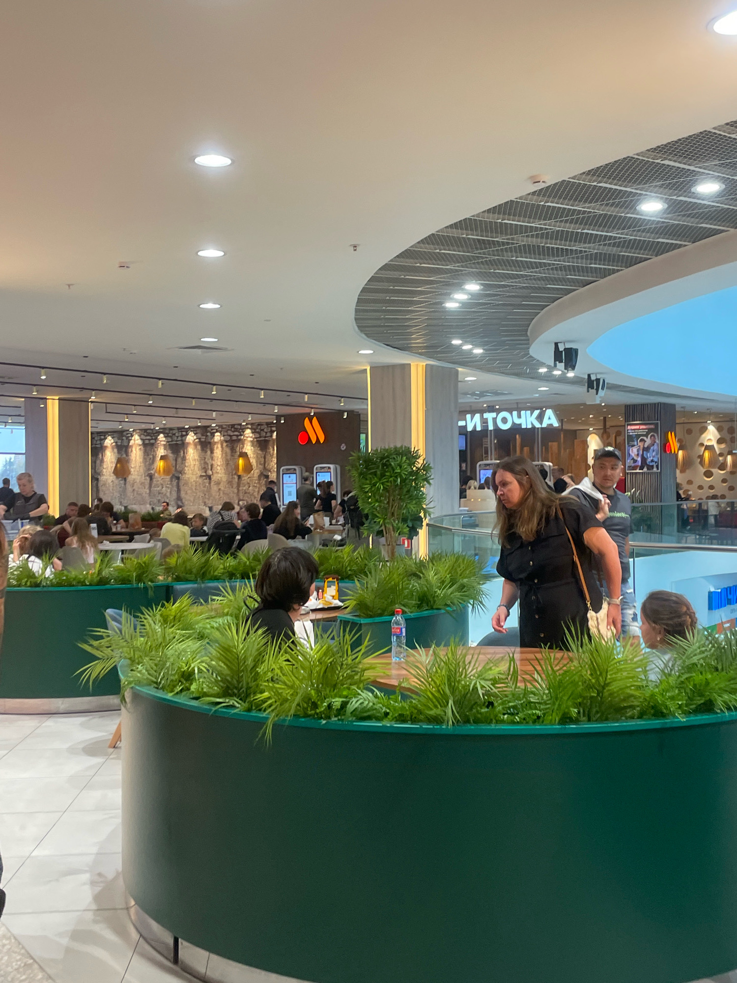 The image depicts a bustling food court within a modern shopping mall in Sochi, Russia. The scene is characterized by an abundance of artificial and natural lighting, with recessed ceiling lights illuminating the space and decorative lamps on the walls of the food establishments. In the foreground, a large, dark green planter filled with lush green foliage divides the seating area. Several individuals are visible, engaged in various activities.
In the middle ground, a woman in a black dress is walking past a table where a man in a grey t-shirt and a baseball cap is standing. Further into the food court, numerous people are seated at tables, some eating, others conversing. In the background, the signage for various food vendors is visible, including one with a distinctive orange "M" logo, likely representing McDonald's, and another that reads "И ТОЧКА" (I TOCHKA), which is the current brand for McDonald's in Russia. The architecture of the mall is modern, with curved ceilings and metal grid details. The overall atmosphere suggests a lively and commercial environment.