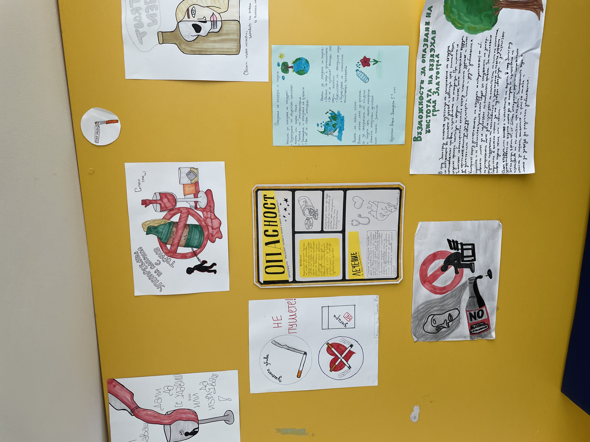 The image shows a yellow wall covered with posters and drawings, mostly related to health and environmental awareness. Topics include dangers of alcohol and drugs, the importance of clean air and water, and anti-smoking messages. There's also a handwritten paper mentioning Zlatograd, Bulgaria, and how to protect the city's air quality. Other messages, such as "Stop Smoking" and "Danger, Narcotics harm the brain," are also displayed.