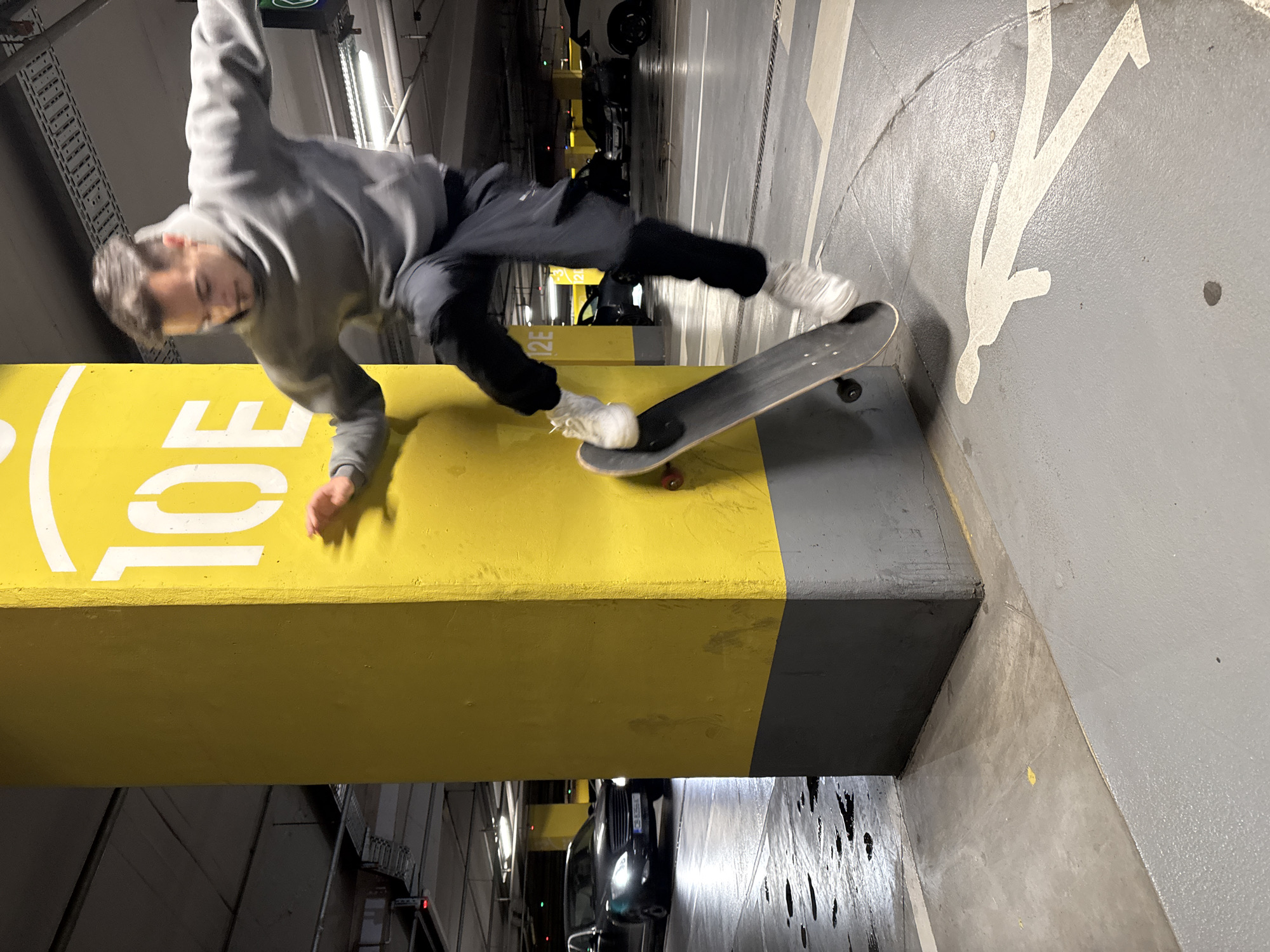 An action shot captures a male skateboarder in mid-air, performing a trick within an underground parking garage in Sofia, Bulgaria. The image is oriented sideways, but if viewed upright, the skateboarder is positioned to the left of the frame, caught in dynamic motion above a prominent yellow and grey concrete barrier or ledge. He wears a grey hooded sweatshirt, dark trousers, and white sneakers. His left arm is extended downwards, appearing to touch the yellow surface, while his right arm is bent upwards. His body is blurred, conveying rapid movement and focus on the trick.
A black skateboard with white trucks and wheels (one wheel showing a red hub) is balanced precariously on the edge of the yellow barrier, suggesting a grind or slide maneuver. The barrier itself is a substantial concrete structure, painted bright yellow on its upper section, transitioning to grey below. White numbers and letters, "10" and a partial "E", are visible on the yellow surface of the barrier.
The setting is clearly a parking facility, characterized by a grey concrete floor with white markings, including a clear pictogram of a walking person. Bright fluorescent lighting fixtures illuminate the ceiling, and metal infrastructure is visible throughout. In the background and below the barrier, parked or passing dark-colored cars are visible, reflecting the artificial light. The overall atmosphere is industrial and functional, serving as an unconventional arena for skateboarding. The motion blur on the skateboarder indicates a moment of intense activity.