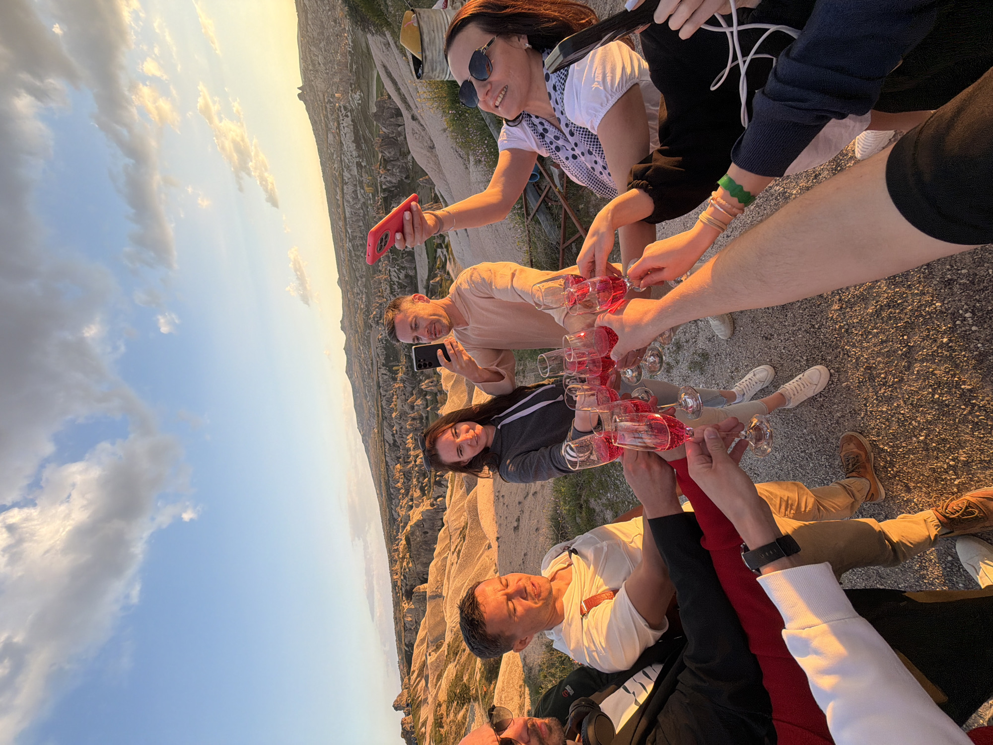 This image depicts a joyful group of seven people gathered for a toast outdoors, set against the distinctive geological formations of Göreme, Cappadocia, Turkey, during the golden hour. The scene is illuminated by the warm, soft light of the setting sun, casting long shadows and creating a golden glow on the horizon, while the sky above is a clear blue with scattered clouds.
The central activity involves the group raising glasses filled with a reddish-pink sparkling beverage, suggesting a celebration or a special moment. Most individuals are smiling and appear happy, contributing to a festive atmosphere. A woman on the right, wearing sunglasses and a white top with black polka dots, holds a red smartphone, possibly capturing the event. Below her, a man in a light brown shirt also holds a black smartphone, seemingly taking photos or videos. Another woman in a grey hoodie smiles directly at the camera. Other participants are visible through their hands holding glasses and partial figures, indicating a larger group sharing the experience.
The unique landscape of eroded rock formations, known as "fairy chimneys," forms the natural backdrop, confirming the location in Cappadocia. The ground beneath the group is gravelly and uneven. There is no visible text in the image. The overall impression is one of camaraderie and enjoyment in a stunning natural setting at sunset.
