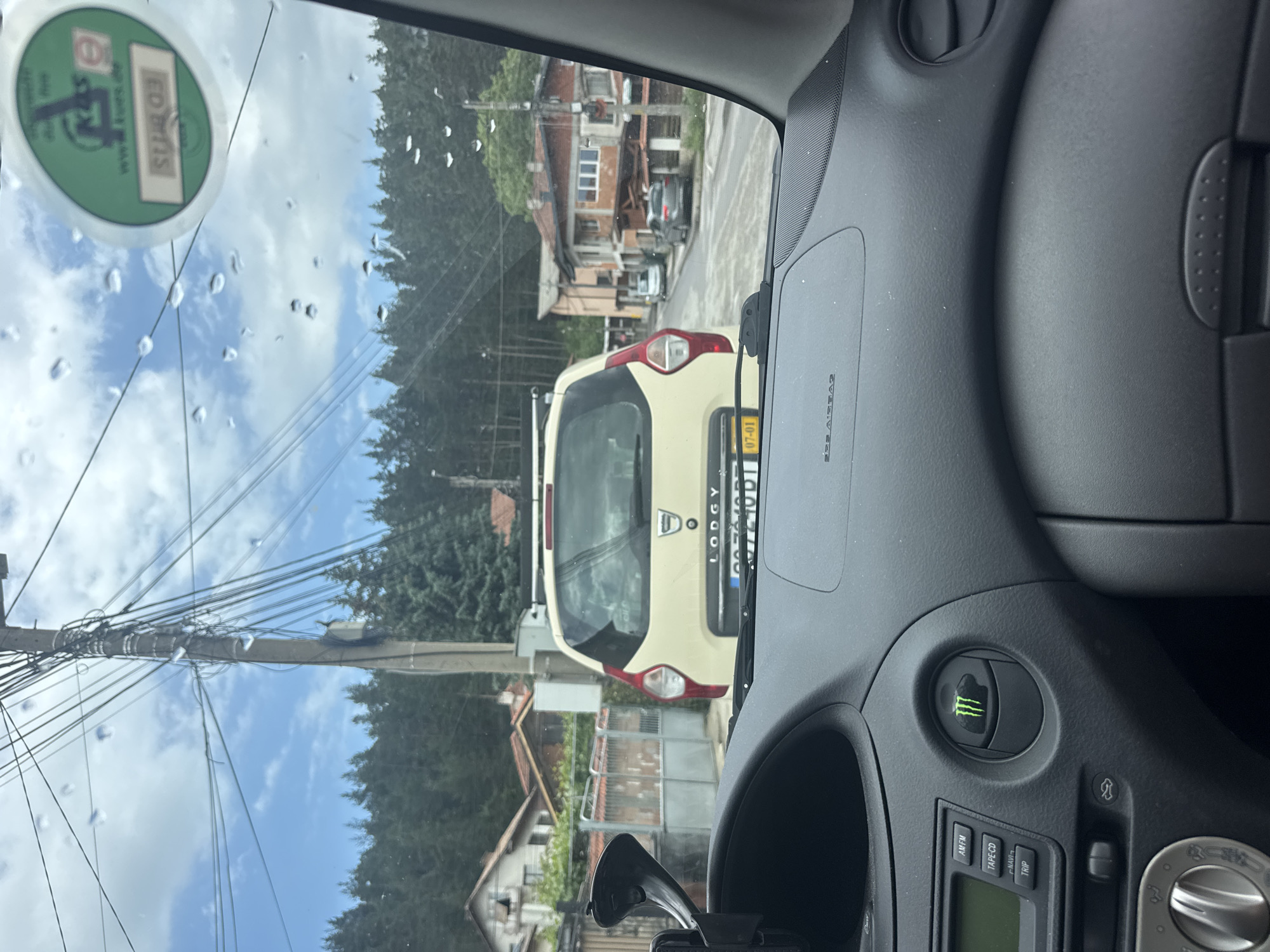 The image depicts a first-person view from inside a vehicle, looking through a rain-splattered windshield at a street scene in Samokov, Bulgaria. The car's dashboard, partially visible in the foreground, features a media player with buttons labeled "AM/FM," "TAPE-CD," "NAVI," and "TRIP," and an air vent customized with a glowing green Monster Energy drink logo sticker. A black suction cup mount is affixed to the windshield on the left.
Outside the vehicle, a light-colored (cream or pale yellow) Dacia Lodgy minivan, equipped with a roof rack, is stopped directly ahead, viewed from its rear. The Dacia's license plate is partially visible, displaying "CA" (indicating Sofia registration in Bulgaria) and the numbers "07-01" on a yellow stripe. A small "elf" sticker is present on its rear window.
The street is paved and appears slightly wet. A prominent utility pole stands in the center-left, heavily laden with a dense, interwoven network of black electrical and communication cables. Buildings line both sides of the street. On the left, several residential structures are visible, one with a broken window, behind a metal fence and gate. On the right, further down the road, more residential buildings with tiled roofs are present, some appearing to be in various states of construction or repair. Two additional vehicles, a light-colored sedan and a dark-colored SUV, are parked or moving further down the street on the right.
The background features a dense forest of evergreen trees covering a distant hillside under a partly cloudy blue sky. A round, green European emission sticker (Umweltplakette), partially obscured but showing "www.kues.de" and "SU-03," is visible on the upper right corner of the windshield. The overall environment suggests a semi-urban or rural town setting, consistent with Samokov in Sofia Province, Bulgaria. No individuals are visible.