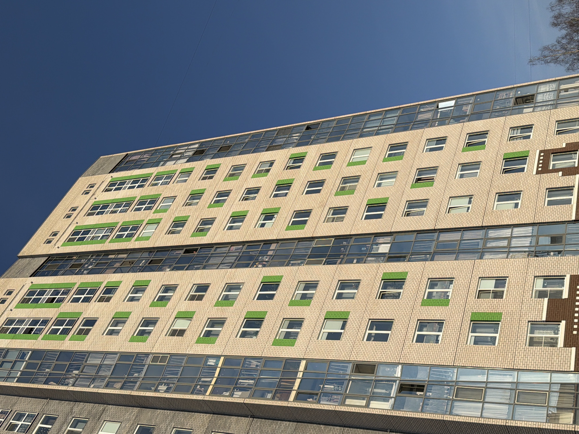The image shows a multistory building in Izhevsk, Russia. The building is beige brick with multiple rows of windows. Some windows have green trim, and the top floor has skylights. The sky is blue and clear. A thin cable stretches diagonally across the upper portion of the image. There are no people visible.