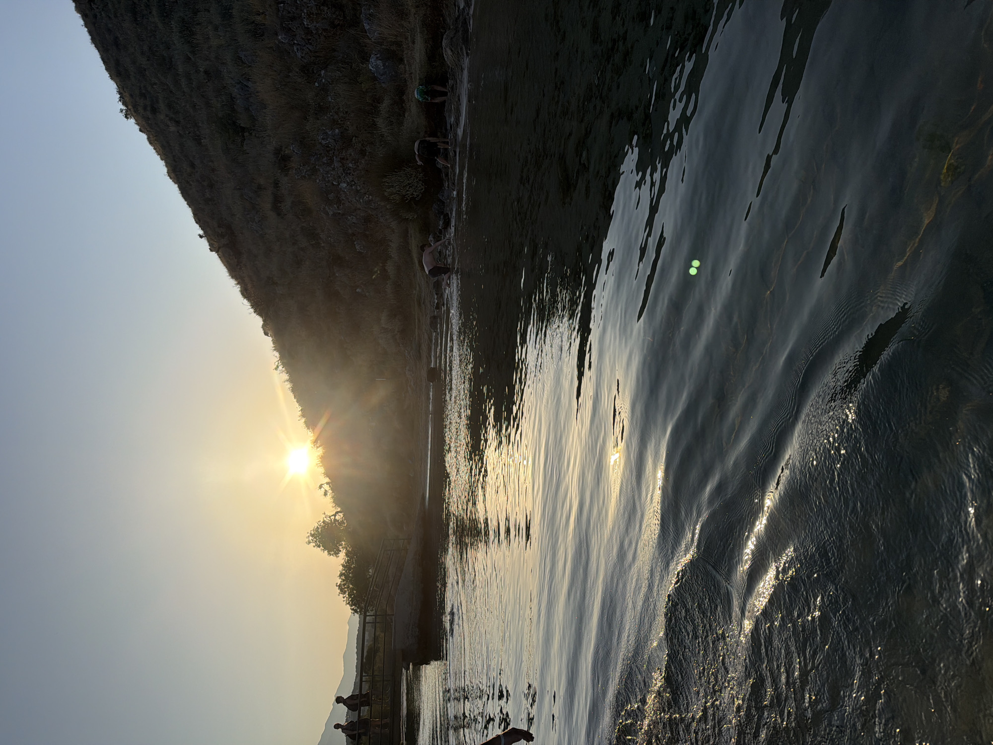 The image depicts an outdoor scene at sunset, featuring a body of water in the foreground and a grassy hillside on the right. The sun is low on the horizon, casting a warm glow and creating sun flares. Several people are present in the scene. On the left, two individuals stand on a walkway or bridge. In the water, closer to the shore, three individuals are bent over, appearing to interact with the water or the shoreline. The water itself is calm with ripples, reflecting the light. The overall environment suggests a natural setting, possibly a lake or riverbank, during the golden hour. There is no identifiable information within the image to confirm a location in Georgioupoli, Greece.