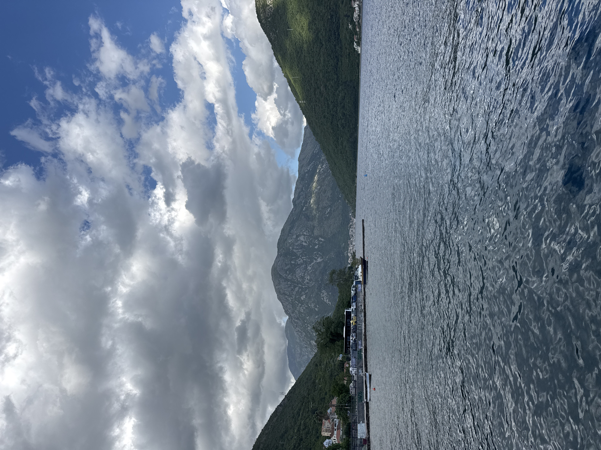 The image shows a waterscape in Đurići, Montenegro. Mountains rise on either side of the water, and a pier extends from the shore into the water. Several boats are docked at the pier. Clouds fill the sky.