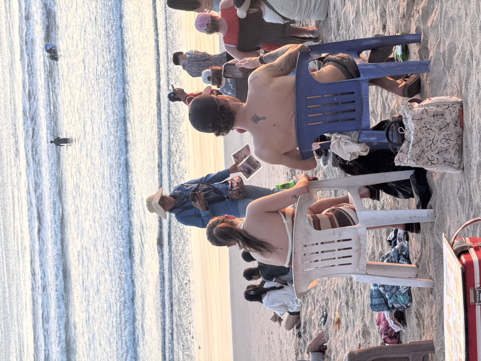A group of people are gathered on the sandy shore of Kuta Beach in Indonesia. The sun is setting, casting a warm glow on the scene. In the foreground, a man with a tattoo on his back is sitting on a blue plastic chair, holding a smartphone. To his left, a woman in a striped top sits on a white plastic chair, holding a green bottle. Several other people are scattered across the beach, some sitting on the sand, others standing. In the background, waves roll in from the ocean, and a few people are in the water. Beach towels and bags are strewn on the sand.