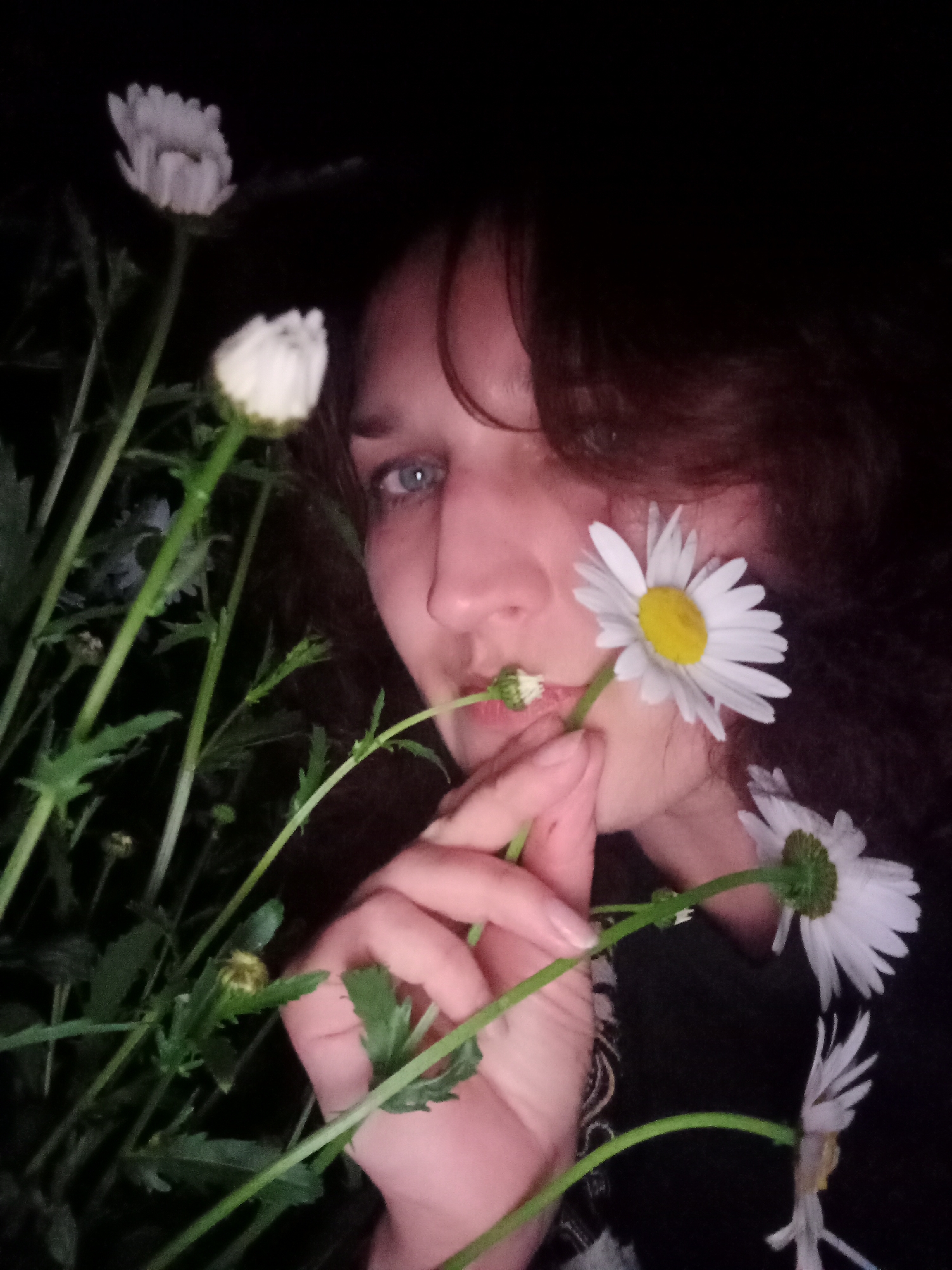 A low-light, close-up shot depicts a person with light skin, brown wavy hair, and blue eyes, holding a cluster of daisy flowers. The person's face is partially obscured by the flowers. The daisies are white with yellow centers, and some are budding. The background is dark, with minimal definition, suggestive of an outdoor setting at night. The location is Babyn, Ukraine.