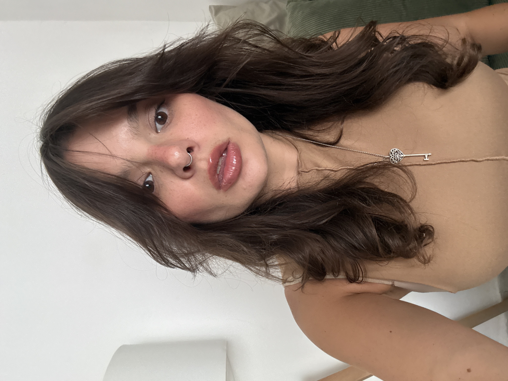 A close-up selfie, taken from a low, upward angle, features a young woman with long, dark brown, wavy hair framing her face. She looks directly into the camera with dark brown eyes and a neutral, direct expression. Her lips are glossy, and she wears a silver hoop piercing in her nose. Her skin has a natural appearance, possibly with light freckles.
She is wearing a light beige or nude-colored strap top, visible across her chest and shoulder. A thin silver chain hangs around her neck, featuring a distinctive key-shaped pendant with an ornate, heart-like design on its head.
The background includes a plain white wall or surface in the lower and left portions of the image, contrasting with a dark green, textured (possibly ribbed or corduroy) pillow or cushion visible behind her head and upper right shoulder. A subtle hint of light-toned furniture can be seen at the bottom right edge. The lighting is soft and even, suggesting an indoor setting. No text is visible. The image is set in Tel-Aviv, Israel.