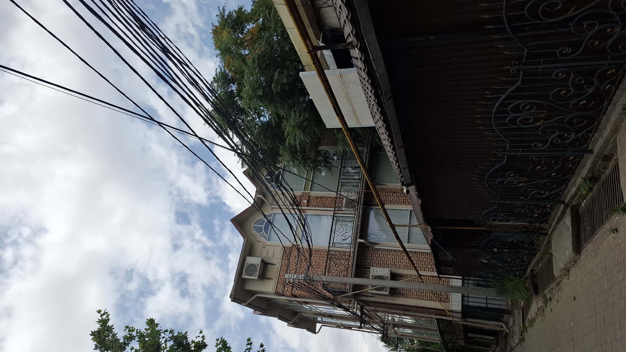 The image depicts a multi-story brick building with large windows and balconies, situated in a city environment. Overhead, a tangle of black electrical wires crisscrosses the sky, obscuring parts of the building and a lush green tree. A dark, ornate metal fence runs along the foreground, bordering a paved walkway or street with drainage grates. The sky above is partly cloudy. The building features air conditioning units mounted on its exterior. The overall scene suggests an urban setting, possibly a residential or commercial area. This image was taken on Sat, 16 Jul 2022, in Shymkent, Kazakhstan.