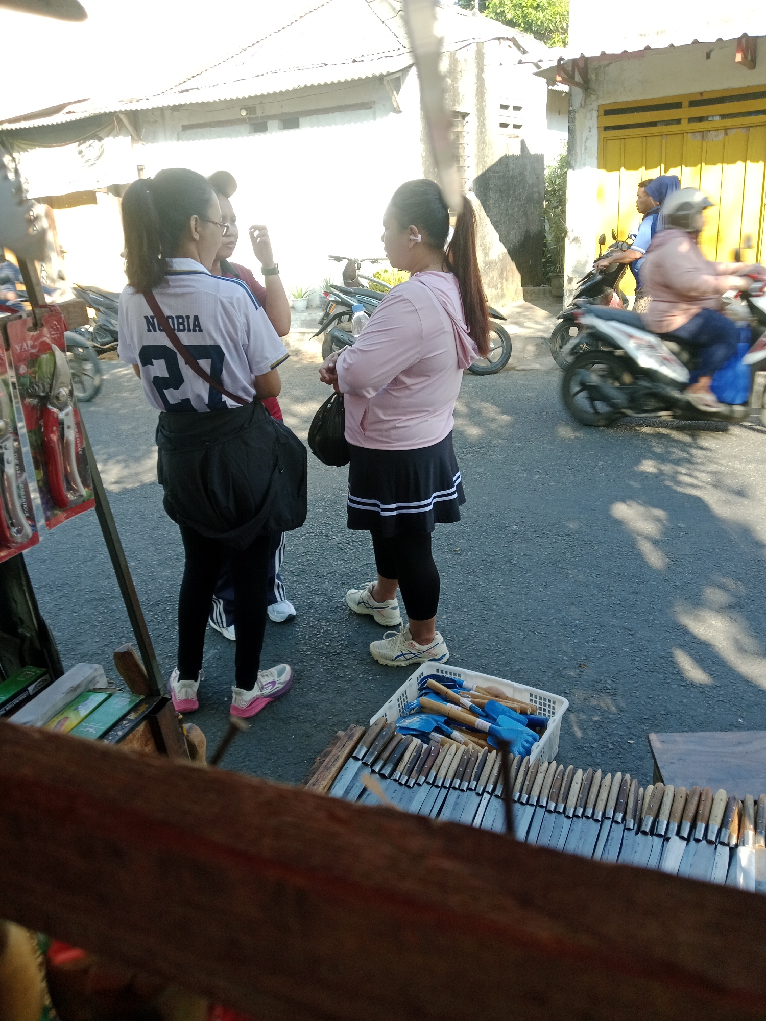 In Kediri City, Indonesia, on a sunlit street, three individuals are present near a roadside stall. A woman in the foreground is wearing a white t-shirt with "NOBIA" and "27" printed on it, black leggings, and white and pink sneakers. She has a black bag slung over her shoulder. Next to her, another woman wears a pink hooded jacket, a black skirt with white stripes, black leggings, and white sneakers. She is holding a black bag. A third person, partially obscured, is also standing nearby. In the background, a yellow building with a garage door and a corrugated metal roof are visible. Several motorcycles are parked or in motion on the street. In the foreground, a stall displays gardening tools, including small shovels and trowels in a white basket, and a row of knives with wooden handles.