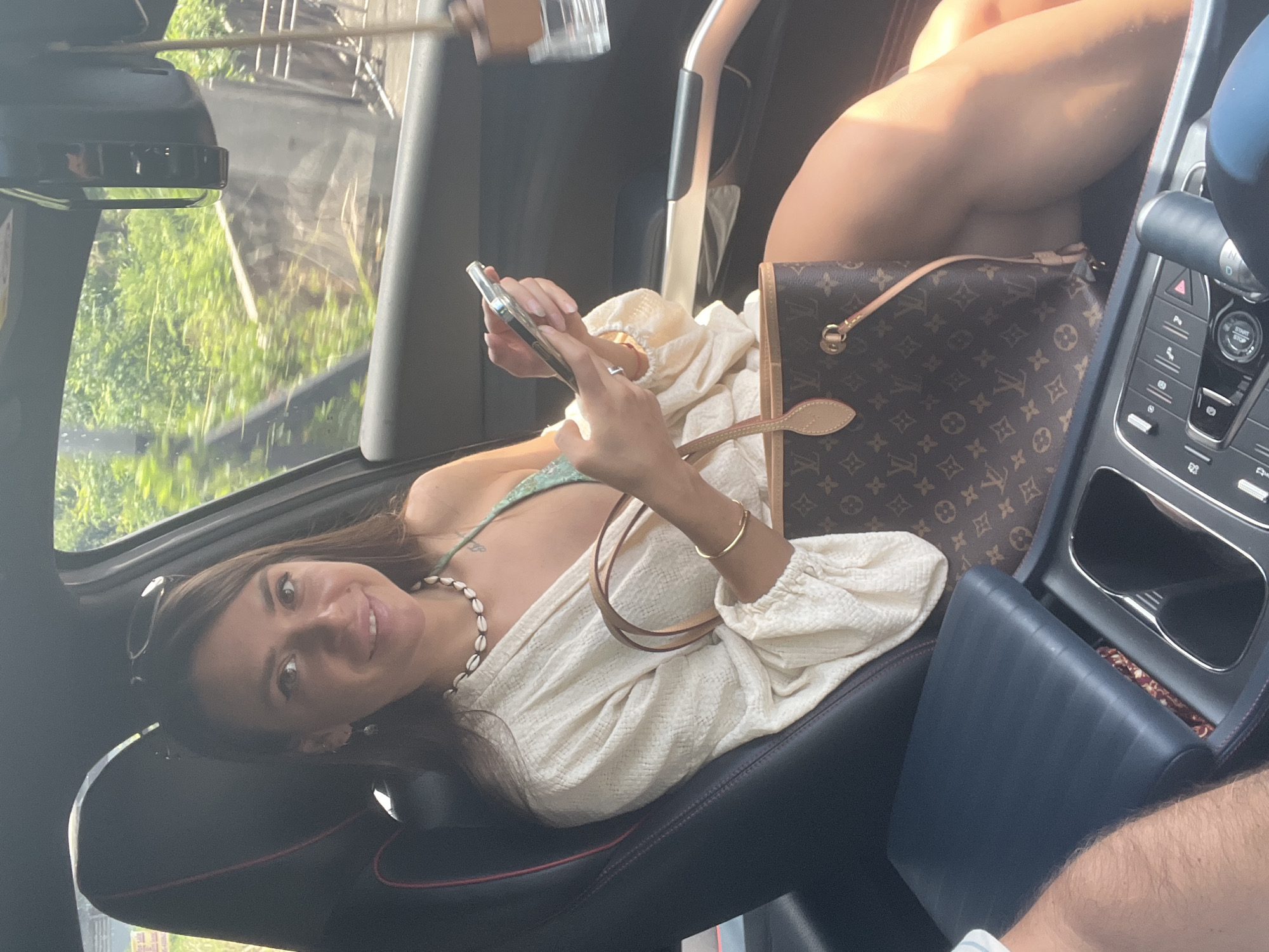 A woman with dark brown hair sits in the passenger seat of a car, holding a smartphone. She is smiling and looking at the camera. She is wearing a cream-colored open-front shirt over a green bikini top, a necklace made of shells, and a gold bracelet. A Louis Vuitton handbag rests on her lap. The interior of the car is visible, including the dashboard and a portion of the center console. Outside the car, lush green foliage suggests a location with abundant vegetation. The scene is well-lit, with natural sunlight filtering into the car. The image does not contain any information that would help to identify the specific location as บ้านบางรักษ์, Thailand.