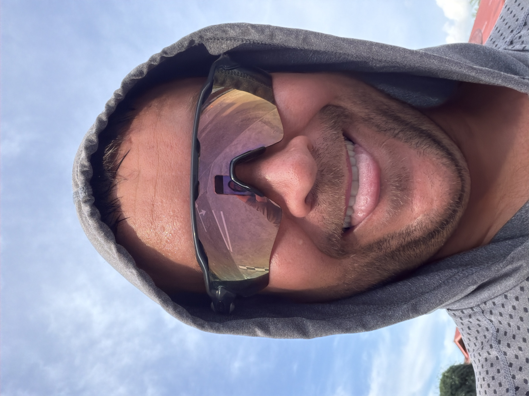 A selfie is taken from a low angle, with a man wearing sunglasses and a grey hooded jacket. The man has stubble on his face and his tongue is sticking out. The sunglasses have a reflection of a road with white lines and a checkered pattern. The background is a bright blue sky with scattered clouds, and some red roofing and greenery are visible at the bottom of the frame, suggesting an outdoor location possibly in Baan Mae Nam, Thailand.