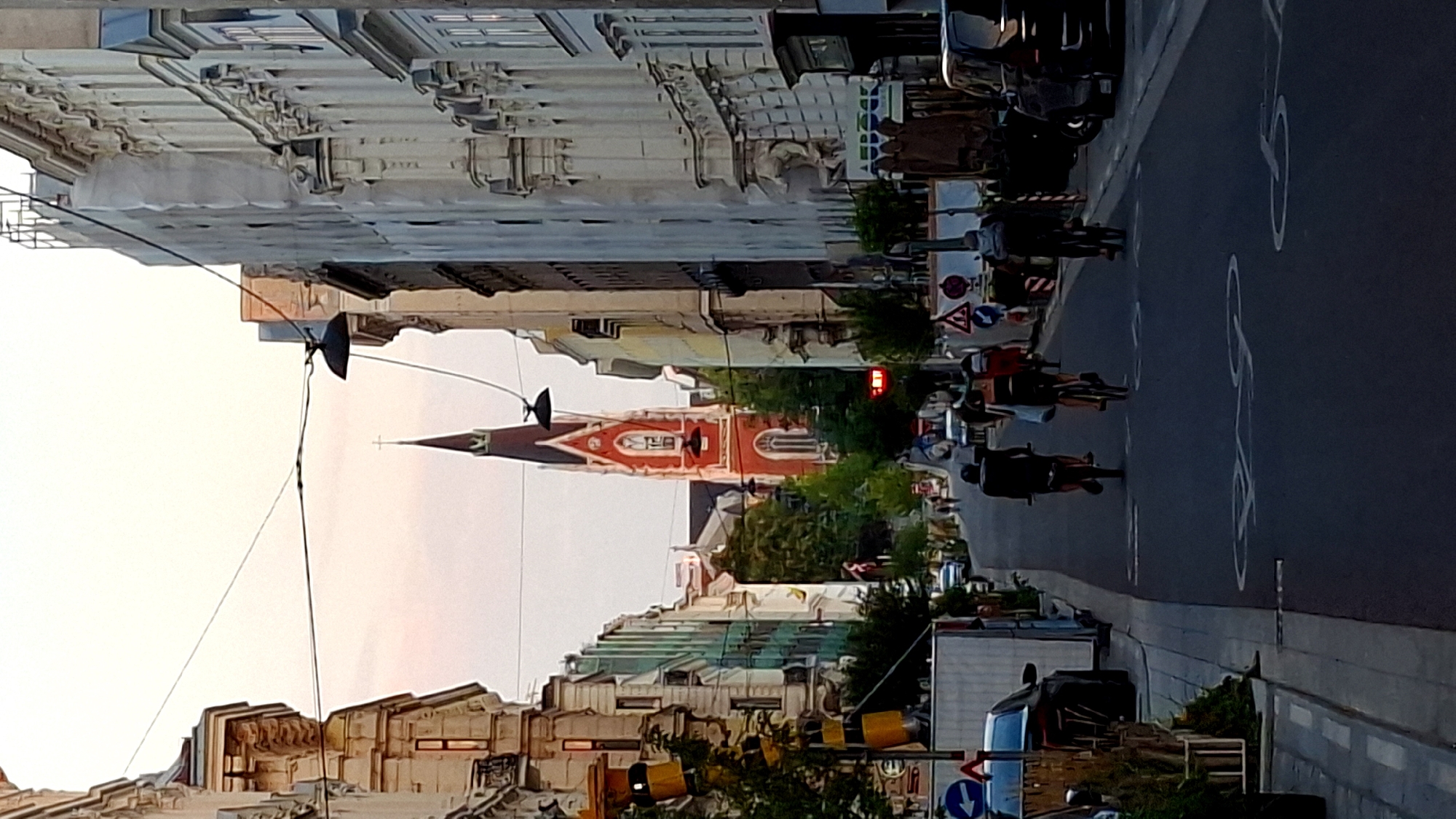The image depicts a street scene in Vienna, Austria, with a prominent red church steeple in the background. The street is lined with buildings on both sides, some of which appear to be under renovation or scaffolding. The sky is a pale pink, suggesting it is either dawn or dusk. Several people are visible on bicycles riding down the street, and there are also cars parked along the sides of the road. Road markings for a bicycle lane are visible on the asphalt. The overall impression is that of an urban environment during a transitional time of day, with a mix of historical architecture and modern urban activity.