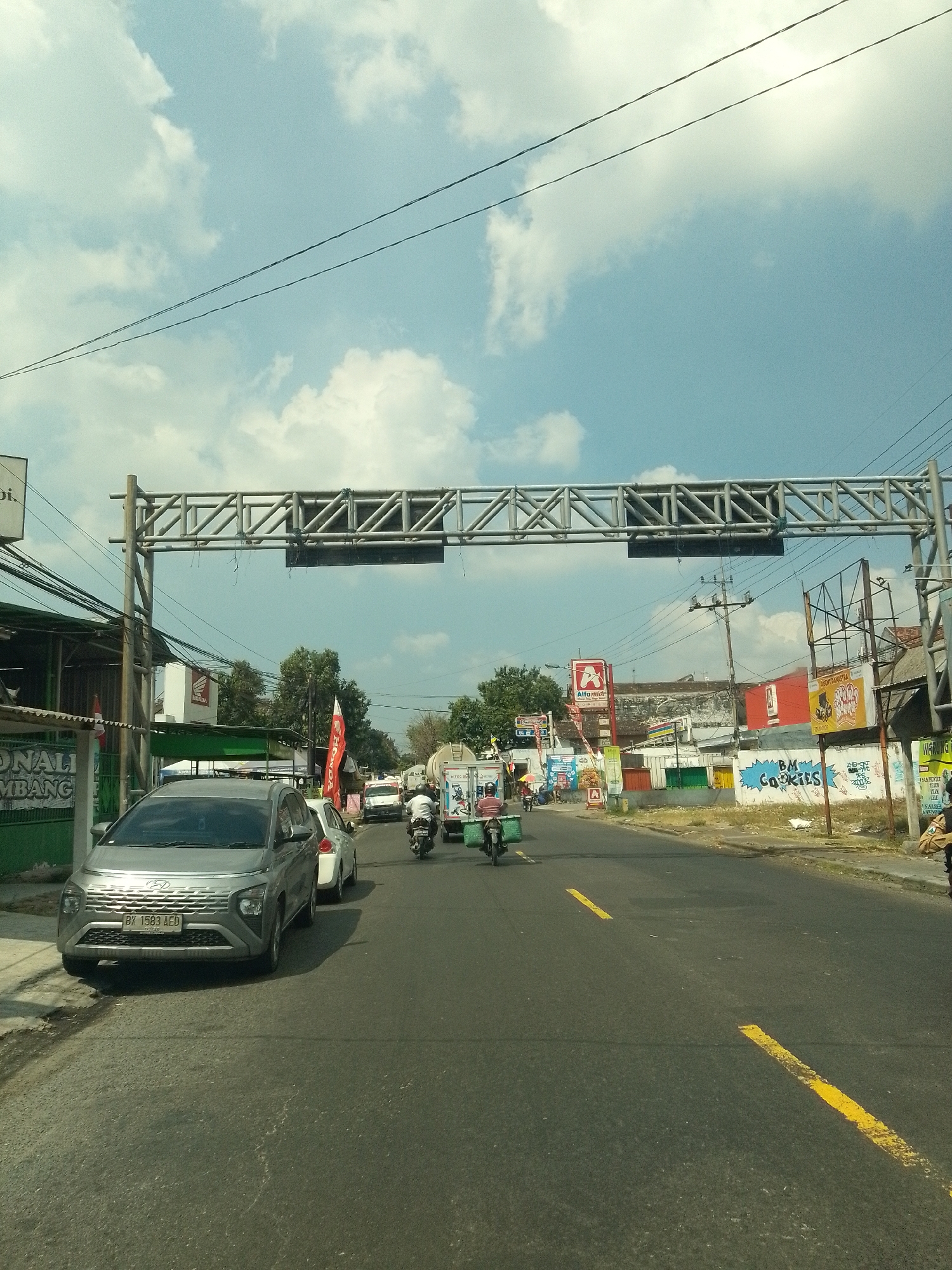 A street scene in Babat, Indonesia, shows a grey SUV with license plate BK 1583 AED stopped at a traffic light. The SUV is in the left lane of a two-lane road. A white sedan is stopped behind the SUV in the same lane. A motorcyclist with a green basket on the back of their motorcycle is to the left of the SUV, also stopped. Several other vehicles, including a truck and other motorcycles, are visible further down the road. The road is paved and has yellow lane markings. On the left side of the road, there are buildings with shops and signs, including a Honda sign. On the right side of the road, there are more buildings, billboards, and utility poles with wires. The sky is blue with scattered white clouds, and a large metal structure, likely a traffic light gantry, spans the road overhead. The scene appears to be during daylight hours.
