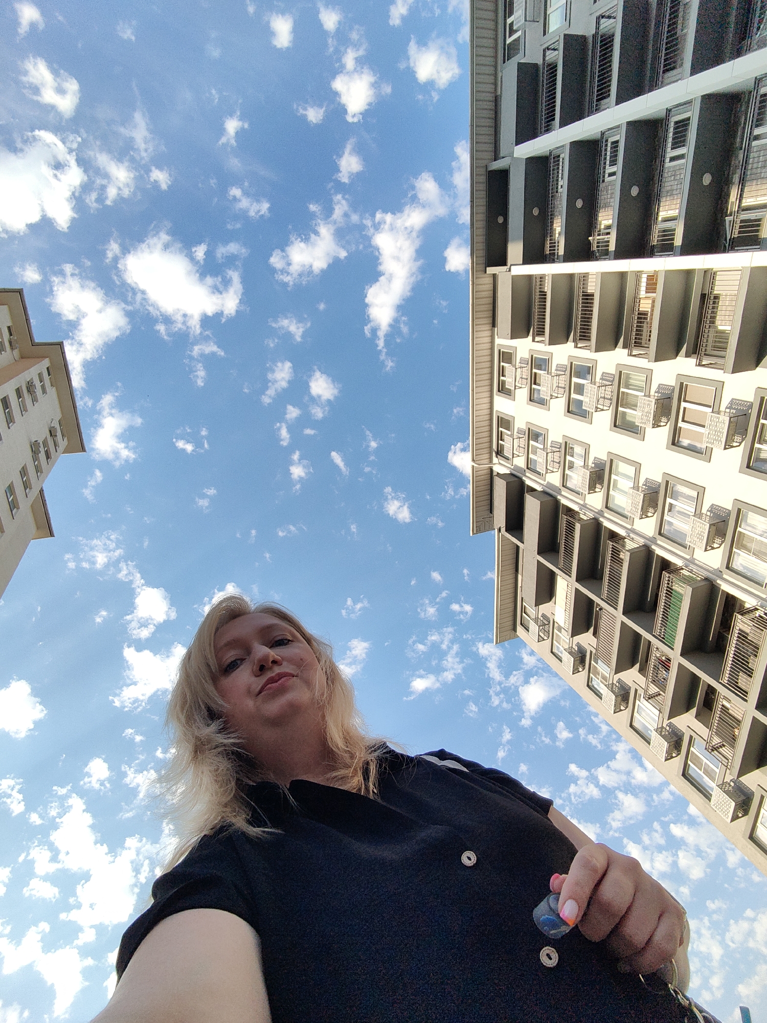 A low-angle, outdoor, wide shot captures a woman taking a selfie from a ground-level perspective. The woman, who has blonde hair and is wearing a black top, is positioned in the lower-center of the frame, looking directly at the camera with a slight smile. Her right hand is visible in the foreground, holding a small blue object. In the background, two modern apartment buildings rise up towards a bright blue sky filled with scattered white clouds. The building on the left is lighter in color, while the building on the right is darker with dark balconies. The scene appears to be set in a city, possibly in Sochi, Russia, given the architectural style and the clear sky.