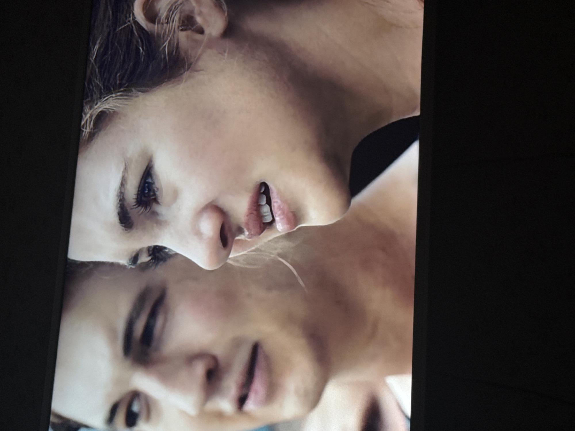 The image shows a close-up shot of two faces, a man and a woman, positioned close together. The woman is on top, with her face slightly angled upwards, showing her eye, nose, and mouth. The man is below her, his face also angled upwards, revealing his eye, nose, and mouth. The lighting is soft, and the background is dark and undefined. The setting cannot be determined from this image, and there is no indication that this image was taken in Mytishchi, Russia.