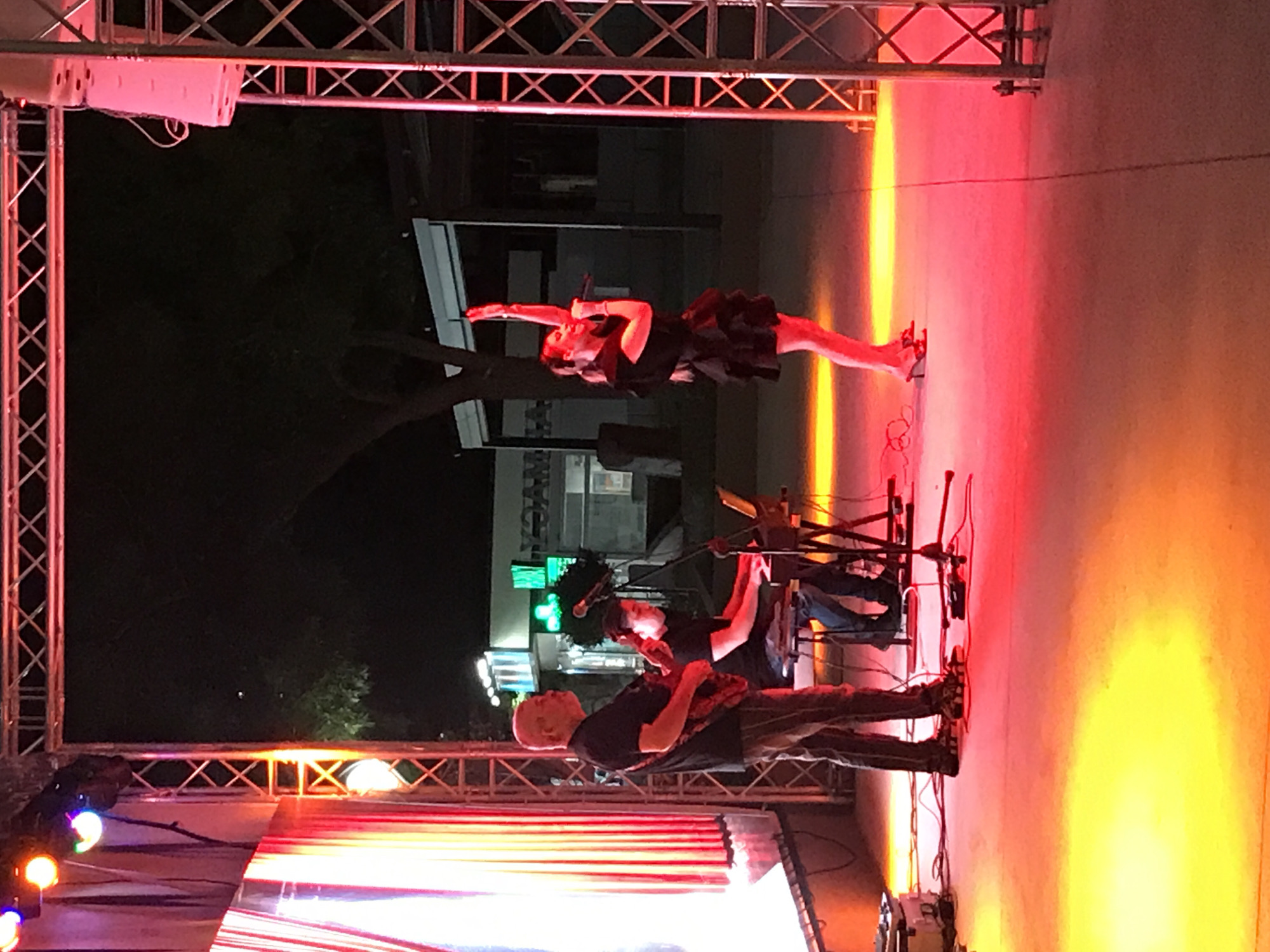 An outdoor evening performance is taking place on a stage. Three individuals are visible: a bald man playing a red electric guitar, a person seated at a keyboard with a laptop, and a woman singing into a microphone while gesturing with her right arm. The stage is illuminated by red and yellow lights, creating a vibrant atmosphere. In the background, a building with illuminated green signage, possibly a pharmacy, is visible, suggesting an urban setting. The overall scene captures a moment of live musical performance.