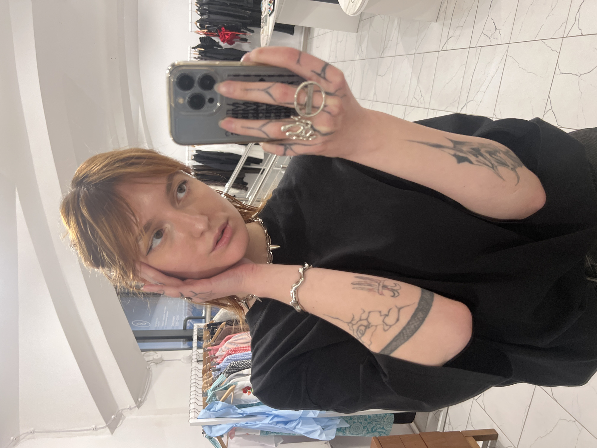 A light-skinned person is taking a selfie indoors. The person has shoulder-length brown hair, and is wearing a black shirt. They are holding a smartphone in their left hand, which is adorned with rings and tattoos. Tattoos are also visible on the right arm. They appear to be resting their face on their hands. The background includes clothing racks with various garments, tile flooring, and a mirror reflecting the scene. The location appears to be a retail store. There is no confirmation that the location is in Saint Petersburg, Russia.