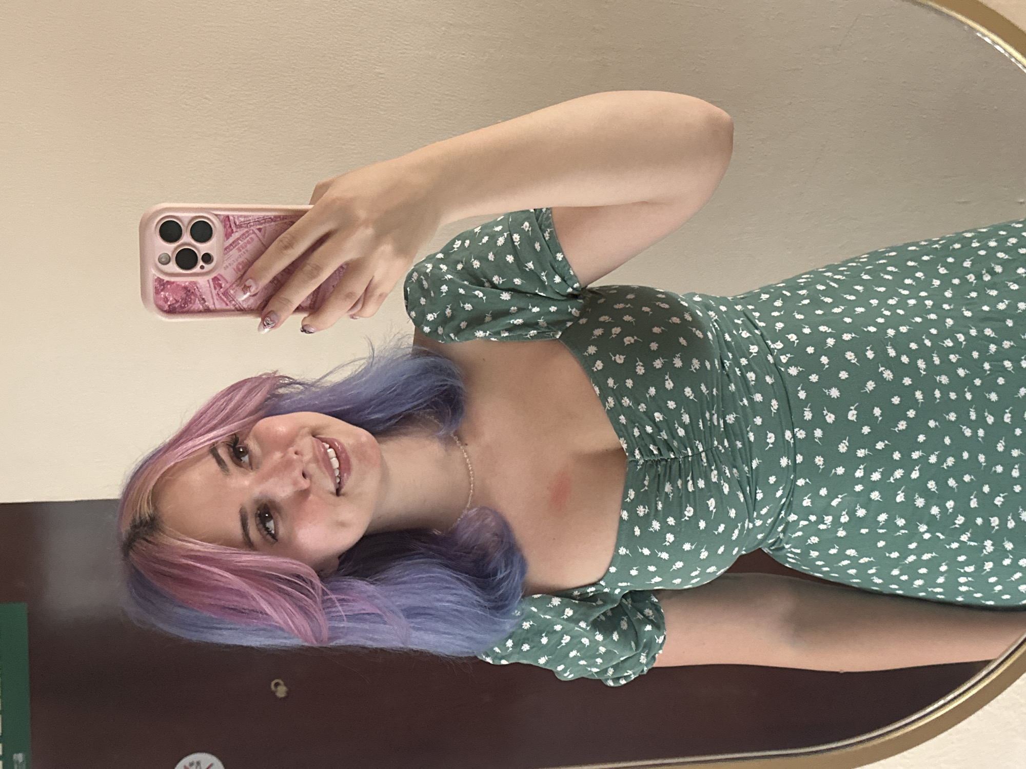 A young woman with multi-colored hair, styled in pink and blue hues, takes a selfie in a mirror. She is wearing a green floral print dress with a sweetheart neckline and puff sleeves. A faint red mark is visible on her chest. She is holding a pink phone case with a camera visible on it. The background appears to be an indoor setting, with a portion of a brown door visible on the left and a beige wall on the right. The overall lighting suggests an indoor environment, possibly a hotel room or a home. There is no indication of the location being in Gijón/Xixón, Spain.