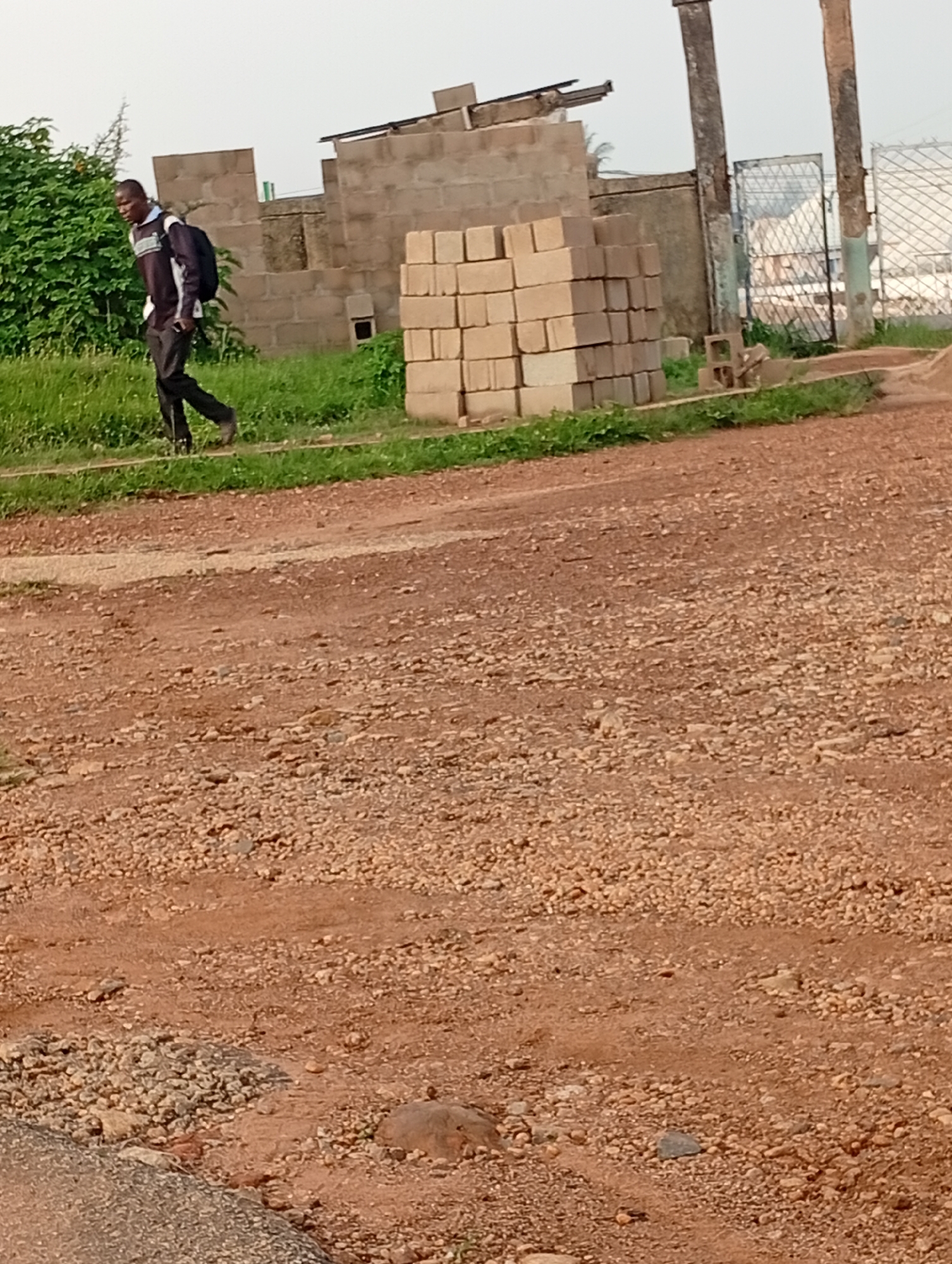 A young Black man walks to the left, his head down, carrying a dark backpack. He is dressed in a black and white striped long-sleeved shirt and dark trousers, with dark shoes. He is walking on a dirt path beside a grassy area. In the background, there is an unfinished brick building with stacks of bricks. Beyond that, a metal fence is visible. The scene appears to be in an outdoor, undeveloped area, possibly on the outskirts of Rafin Par, Nigeria. The ground is covered in red dirt and small stones. The sky is overcast.