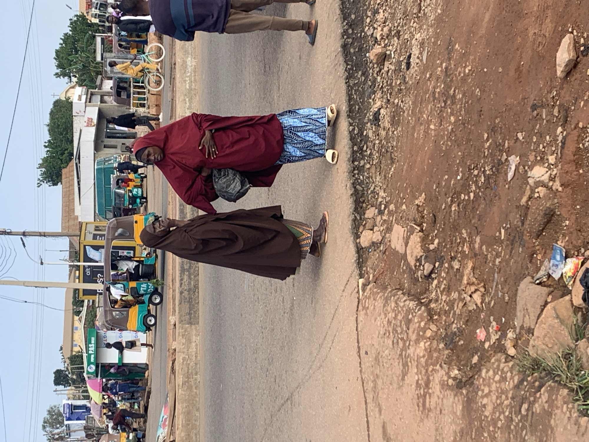 This is a medium shot of a street scene in Gura Top, Nigeria. Two women are standing on a concrete curb. The woman on the left is wearing a brown abaya and a patterned skirt. The woman on the right is wearing a maroon abaya and a blue and white patterned skirt, with white sandals. She is holding a dark bag. Behind them, there is a street with several vehicles, including a yellow and green auto-rickshaw, and a bus. People are walking and standing around. The ground in the foreground is rough and dirty, with some debris. The overall lighting suggests it is daytime.