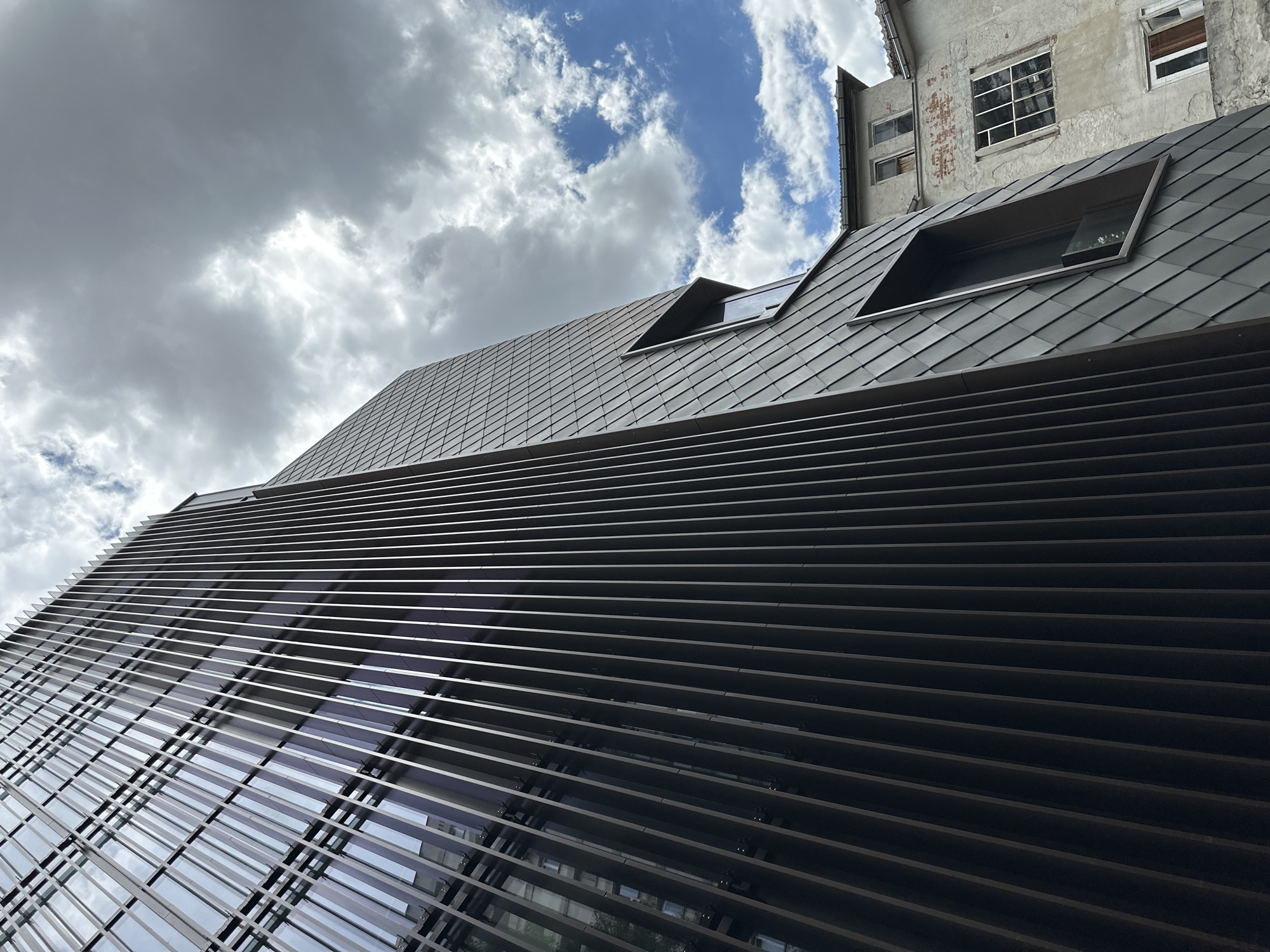 The image shows a modern building with a dark, ribbed facade, contrasted with an older, dilapidated building in the background. The modern building features a glass exterior with vertical metal fins, reflecting the sky and clouds. Its facade is partially covered in dark, diamond-shaped panels, with a prominent, angular window. The older building has a weathered, light-colored exterior, with visible brickwork and multiple windows, some of which appear broken or boarded up. The sky is partly cloudy, with patches of blue visible. The scene is located in Sofia, Bulgaria, and the buildings are situated next to each other, creating a stark contrast between old and new architecture. There are no people or vehicles visible in the image.