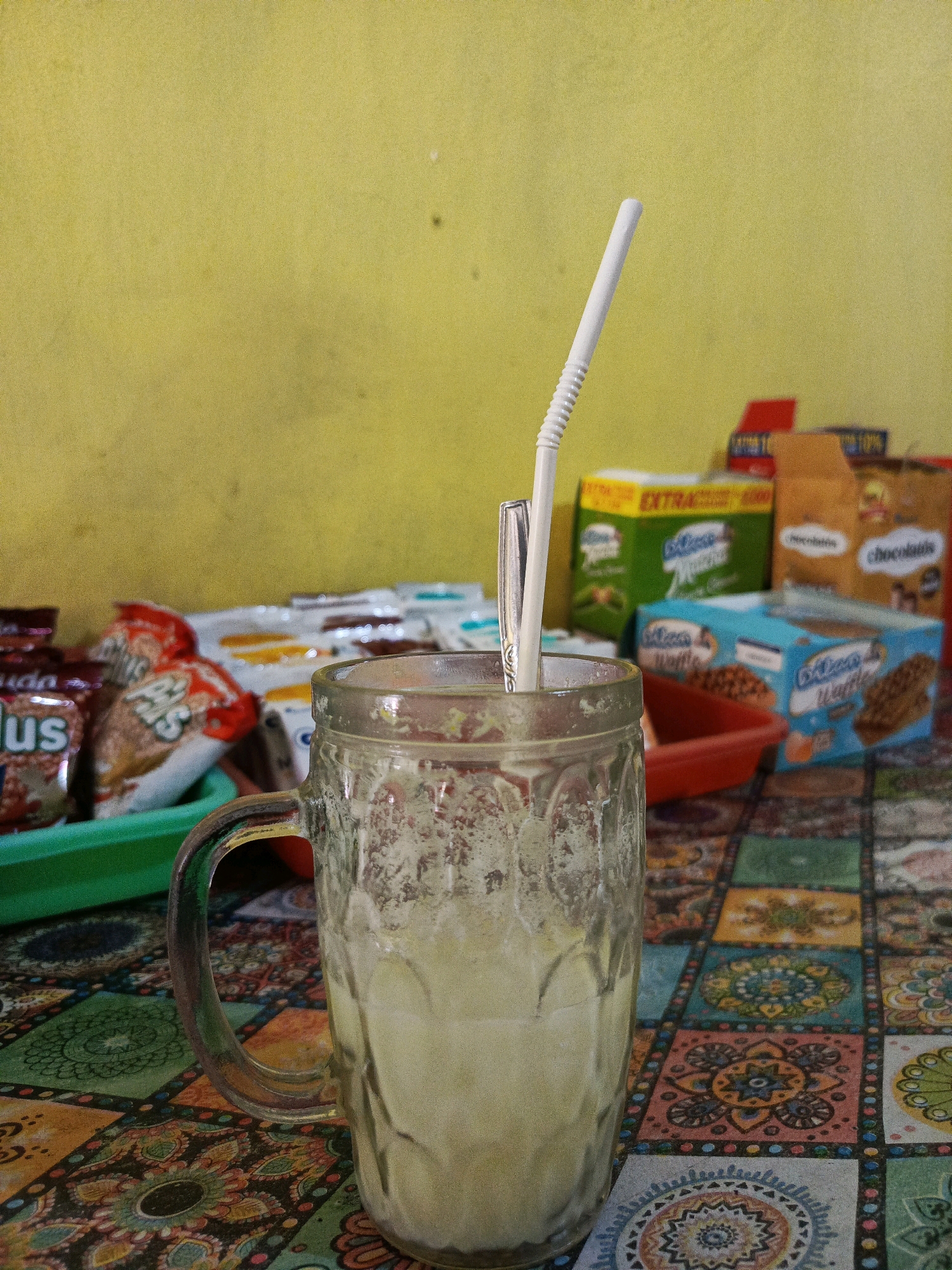 The image depicts a glass of light-colored beverage with a white straw and a metal spoon resting inside, positioned on a table covered with a colorful, patterned tablecloth. In the background, several packaged snacks, including boxes of cookies and other food items, are visible, along with a yellow wall. The overall scene suggests a casual setting, possibly a small shop or a home, where snacks are being consumed with a drink. This image was taken in Pamekasan, Indonesia.