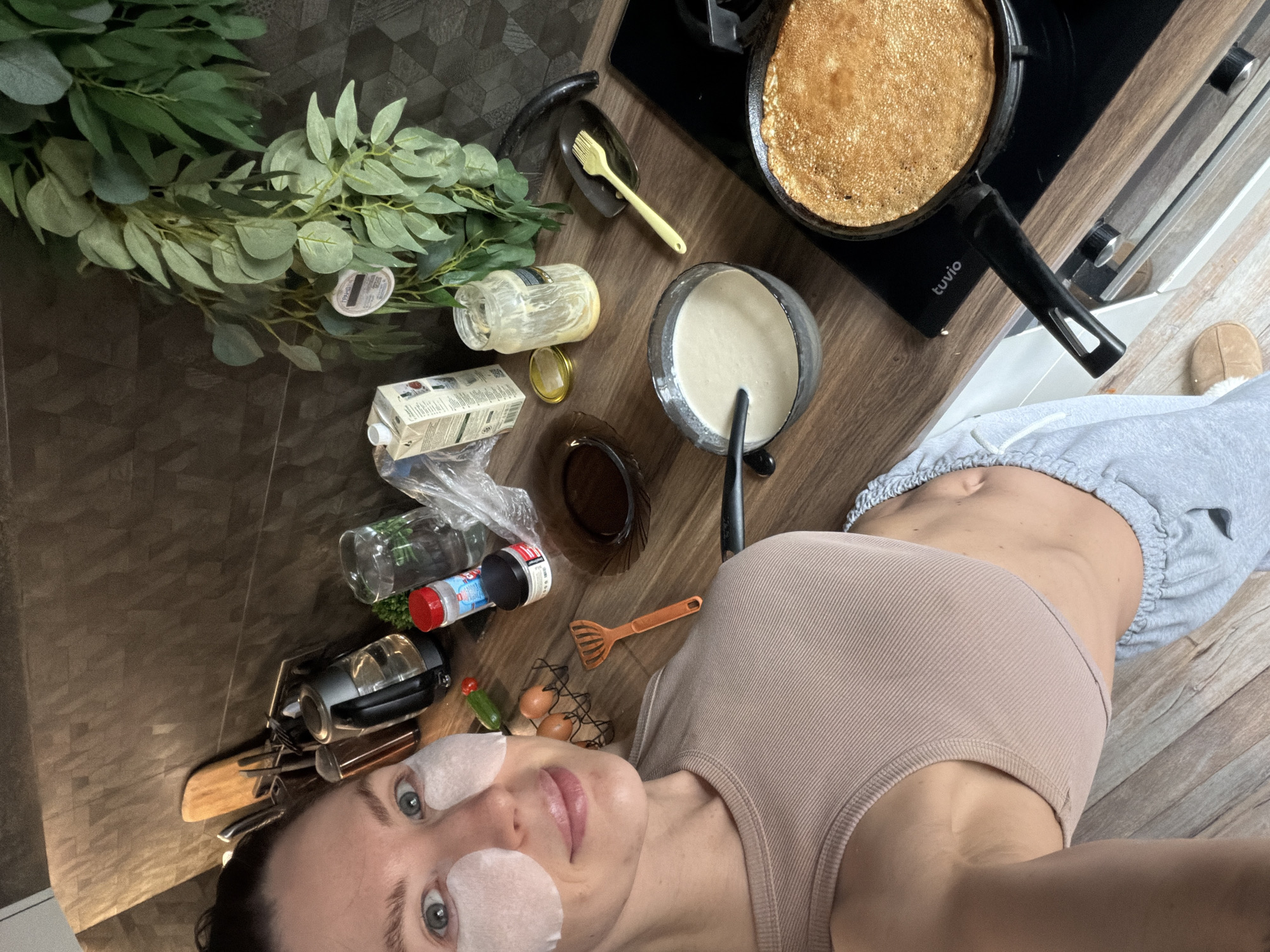 The image shows a woman with eye patches, lying on a wooden floor in what appears to be a kitchen. Adjacent to her, on the floor, are various items, including a frying pan with a pancake inside, a bowl of batter, cooking utensils, eggs, seasonings, empty containers, a carton, and greenery. The frying pan is on a stovetop marked with the brand "tuvio." The specific location is identified as the city of Perm, Russia, based on the information present in the image.