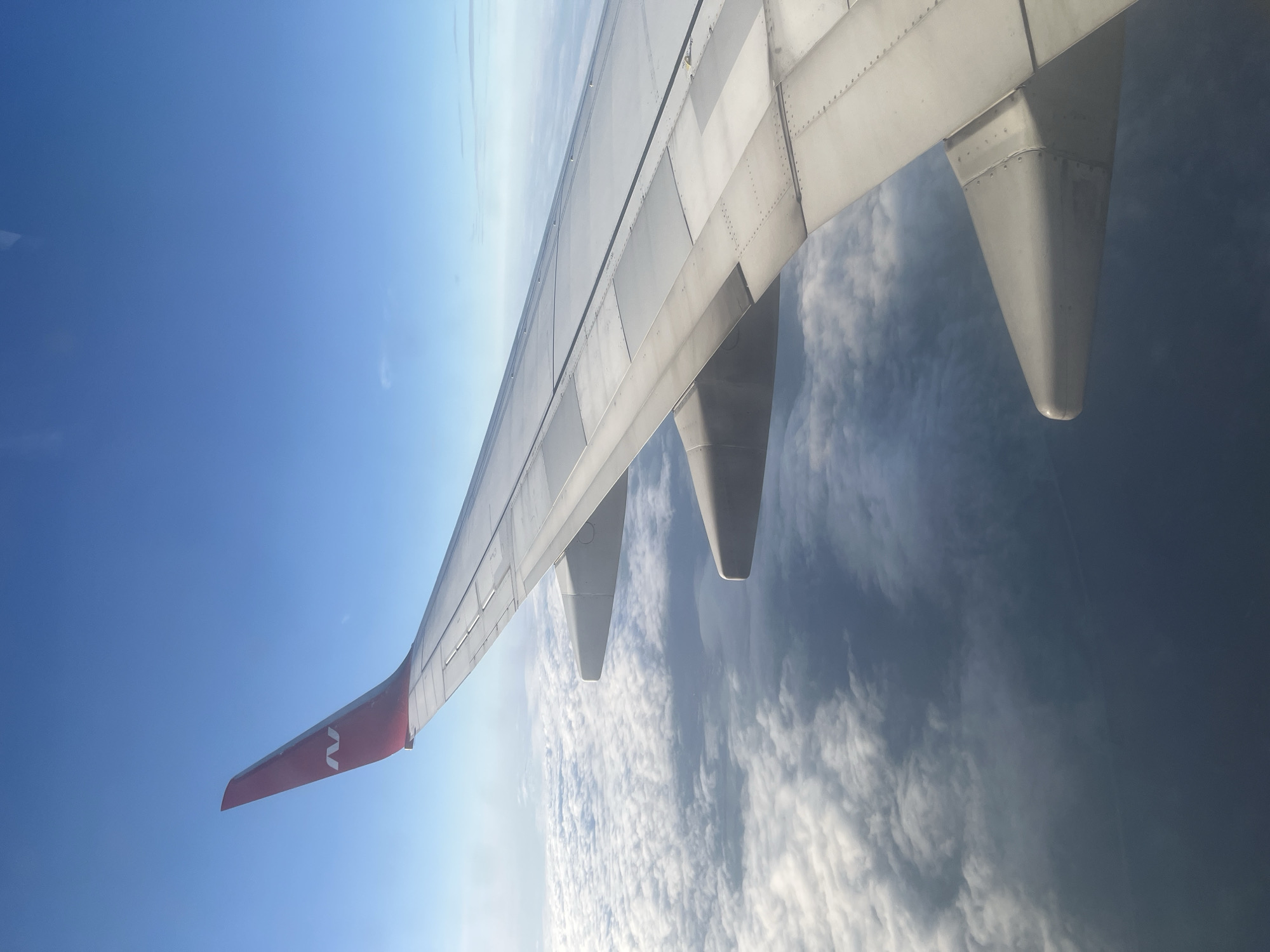 The image is a view from an airplane window, showing a portion of an airplane wing in the foreground. The wing is white with a red tip that has a white logo resembling the letter "N". Below the wing, a vast expanse of white clouds stretches out, interspersed with patches of blue sky peeking through. The sky above is a clear, bright blue. The image does not provide any information or visual cues about the city of Stolbishche, Russia.