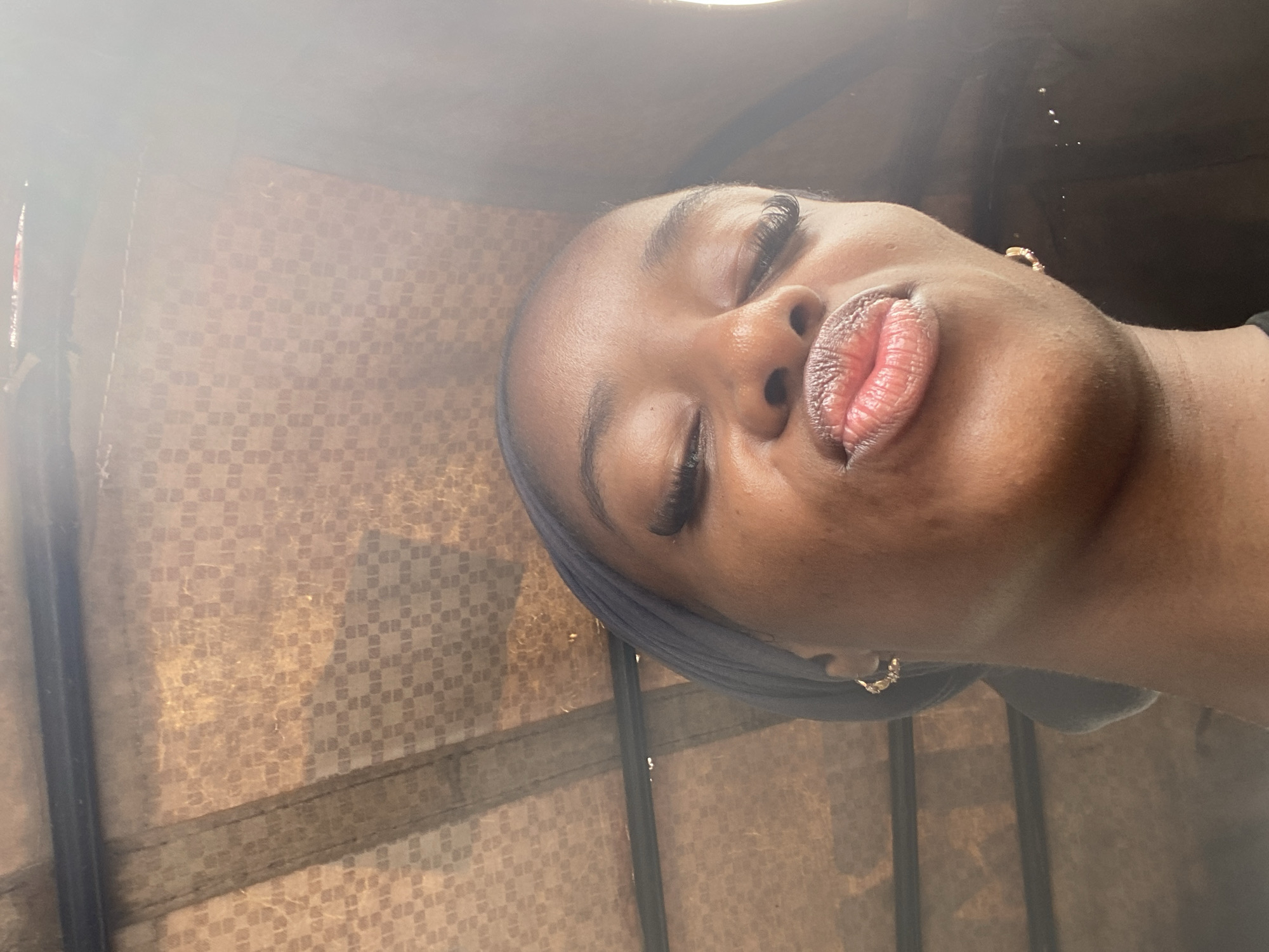 An overhead, close-up selfie shows a young Black woman with closed eyes and pursed lips, wearing a dark headscarf and gold earrings. Her face is angled upwards, with the focus on her features. The background is blurred but appears to be the interior of a vehicle, possibly a bus or van, with a patterned ceiling. The image is likely taken outdoors, given the lighting and the context of being inside a vehicle. The specific location in Jos, Nigeria cannot be determined from the visual information provided.