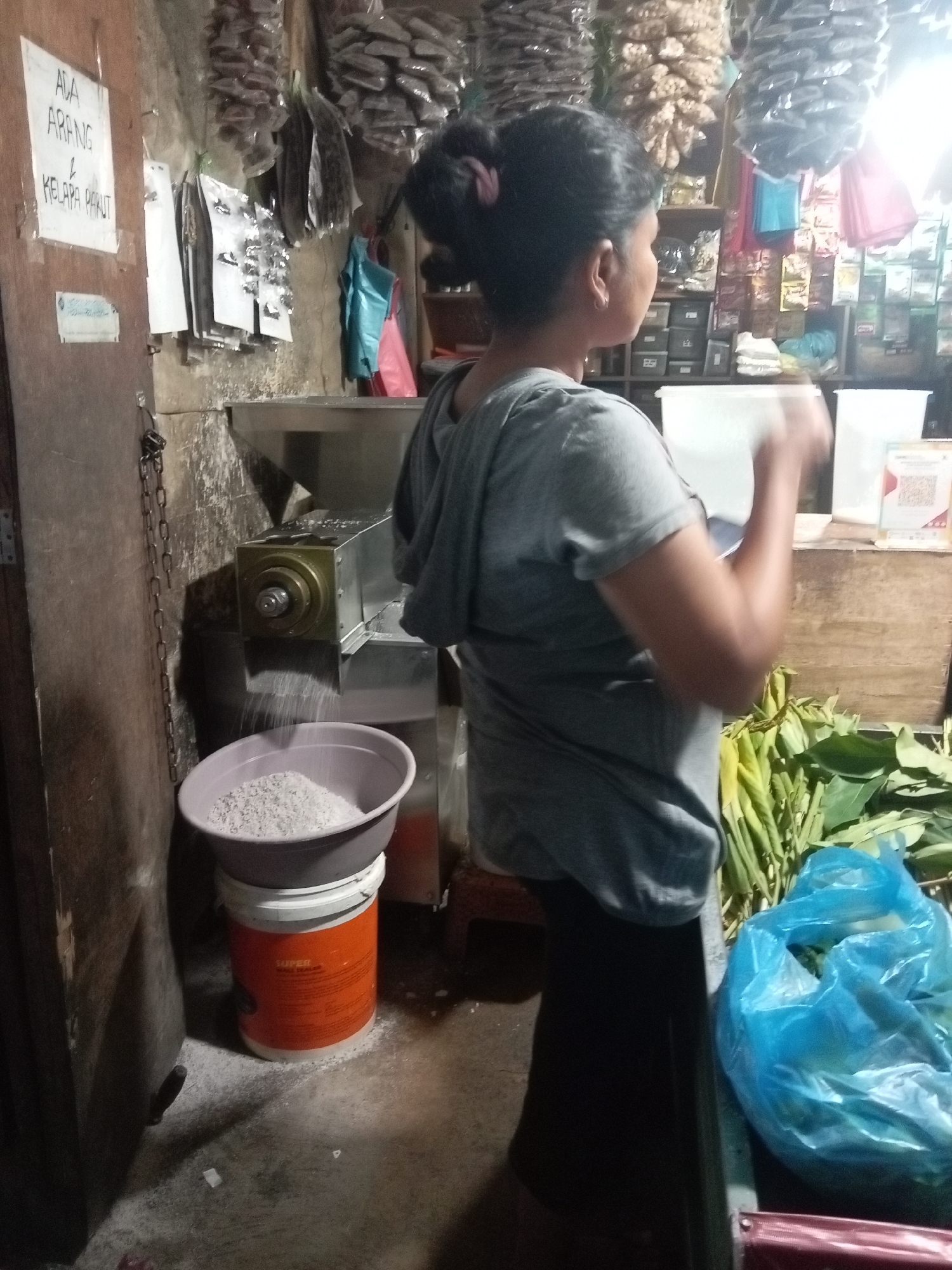 In Batam, Indonesia, a woman with her hair pulled back in a bun, wearing a grey t-shirt and dark pants, operates a machine that appears to be grinding or processing food. The machine, made of metal, is positioned above a grey plastic basin filled with white granular material, likely rice or a similar product. Below the basin sits an orange bucket with some visible text. The woman's hands are blurred, indicating movement as she works.
The background suggests a market or shop setting. Shelves behind the woman are stocked with various items, including what appear to be bags of food or spices, and some blue and pink plastic bags. To the left of the woman, a wooden door has a white sign attached to it with handwritten text in Indonesian. Next to the door, a metal chain hangs. In the foreground and to the right, a pile of green leafy vegetables and a large blue plastic bag are visible. The overall environment appears to be dimly lit with some bright light sources contributing to the illumination.