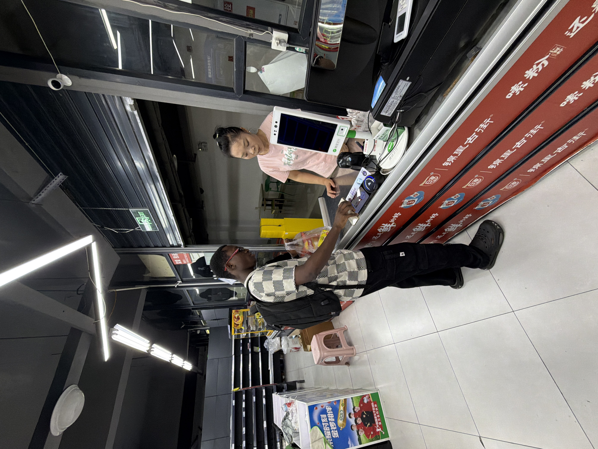 In a supermarket in Jiangjin, China, a Black man wearing glasses, a checkered short-sleeved shirt, and black cargo pants is standing at a checkout counter. He has a black backpack on and is holding a plastic bag with some food items. He appears to be in the process of paying or receiving his purchased items. A woman, possibly a cashier, wearing a pink t-shirt, stands behind the counter, interacting with the man. The counter is equipped with a digital payment terminal, a scanner, and other retail equipment. The background shows shelves, possibly empty, and a security camera on the ceiling. The floor is tiled.