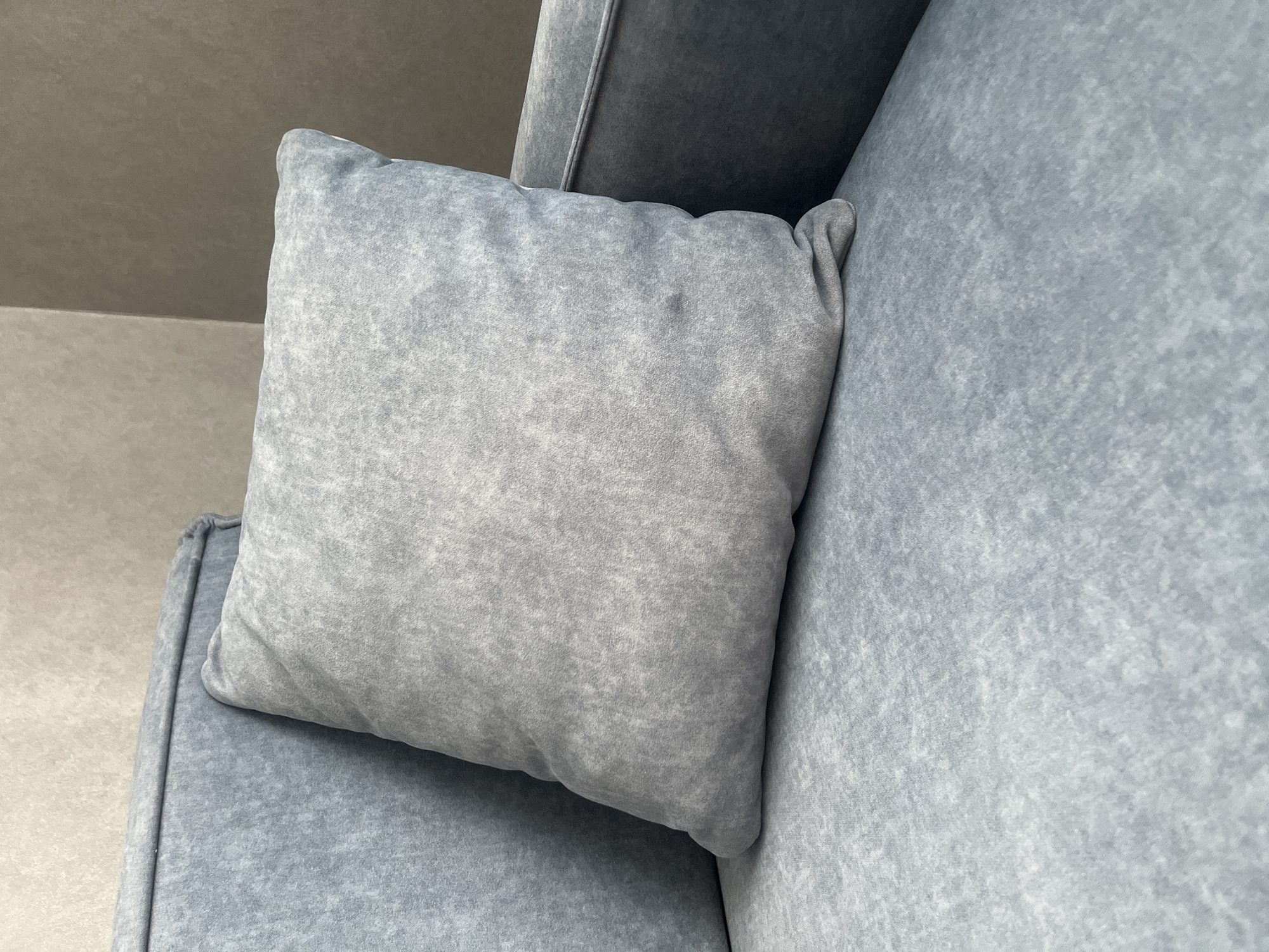 A close-up, eye-level shot focuses on a grey throw pillow resting on a matching grey sofa. The pillow, square in shape, has a textured surface with subtle variations in shading, suggesting a slightly worn or brushed fabric. Its corners are rounded, and the filling appears soft, giving it a relaxed, slightly slumped appearance. The sofa fabric shares the same mottled grey tone, with visible texture and stitching lines. To the left of the pillow, a portion of the sofa's backrest is visible, also in grey. The background consists of a textured wall, transitioning from a lighter beige on the left to a darker brown on the right. There are no people or animals present in the scene. The lighting is soft and diffused, casting subtle shadows. The image does not provide any information that would allow for localization in Rybnitsa, Moldova.