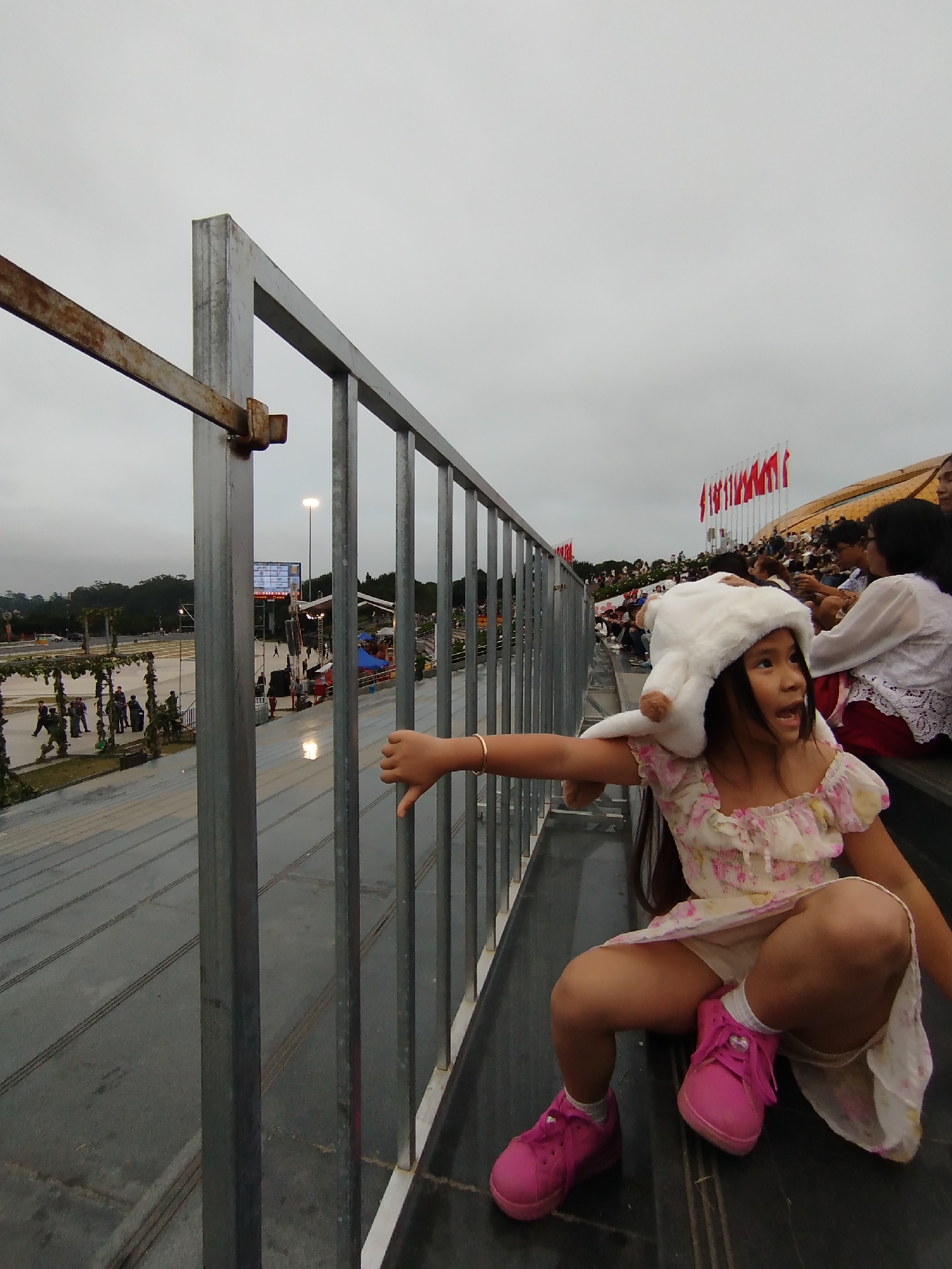 A young girl wearing a fluffy white animal hat and a floral dress sits on bleachers, looking to her right with her mouth open. She has on white socks and bright pink sneakers. Her left arm is extended, with her thumb down, and she wears a gold bracelet on her left wrist. The bleachers are dark gray, and behind the girl, other spectators are visible in the background, some wearing white. A metal railing runs in front of the bleachers.
To the left of the bleachers, a large, paved area is visible, with some greenery and trees in the distance. Several people are gathered in this area, some appearing to be in uniform. A tall light pole with a bright light shines above the scene. In the background, a row of red flags with white symbols is displayed along a curved structure. The sky is overcast, suggesting a cloudy or possibly rainy day. The overall scene appears to be an outdoor event or gathering. There is some visible text on a large screen in the distance, but it is not legible. The location is identified as Da Lat, Vietnam.