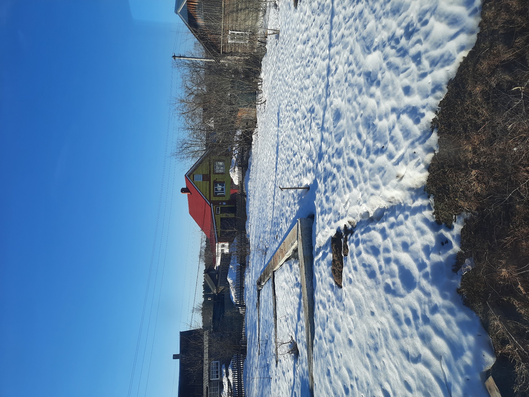 The image depicts a winter scene in what appears to be the Krasnogorskoye area of Russia. A yellowish-green wooden house with a red roof is visible amidst bare trees and a partially snow-covered landscape. A wooden fence runs along part of the property. The ground is covered in snow, with some patches of bare earth and dry vegetation showing through. The sky is clear and bright blue. In the foreground are remnants of a rectangular wooden frame structure, mostly buried under snow. Other buildings are partially visible in the background. Utility poles and wires are present overhead.