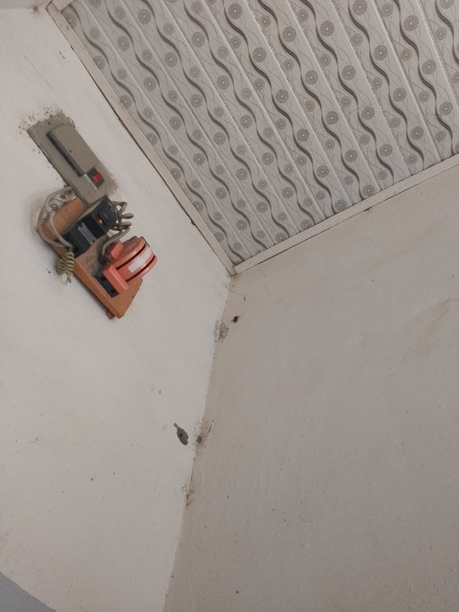 The image depicts an interior scene. The upper portion of the image shows a white ceiling with a decorative pattern of repeating wavy lines and floral motifs in gray. The lower portion shows a textured, off-white wall. On the wall, a gray electrical outlet box is mounted. It contains a black toggle switch and a red button. Below the outlet box, attached to a small wooden base, are electrical components. These include what appears to be a black electrical connector with several wires emerging and an orange device with stacked horizontal lines. The scene is dimly lit, and dust is visible on the electrical components and the wall. There are also small dark spots on the wall that could be marks or insects. No people are visible in the image. The specific location in Jauro Yinu, Nigeria, cannot be determined from the visual information provided.