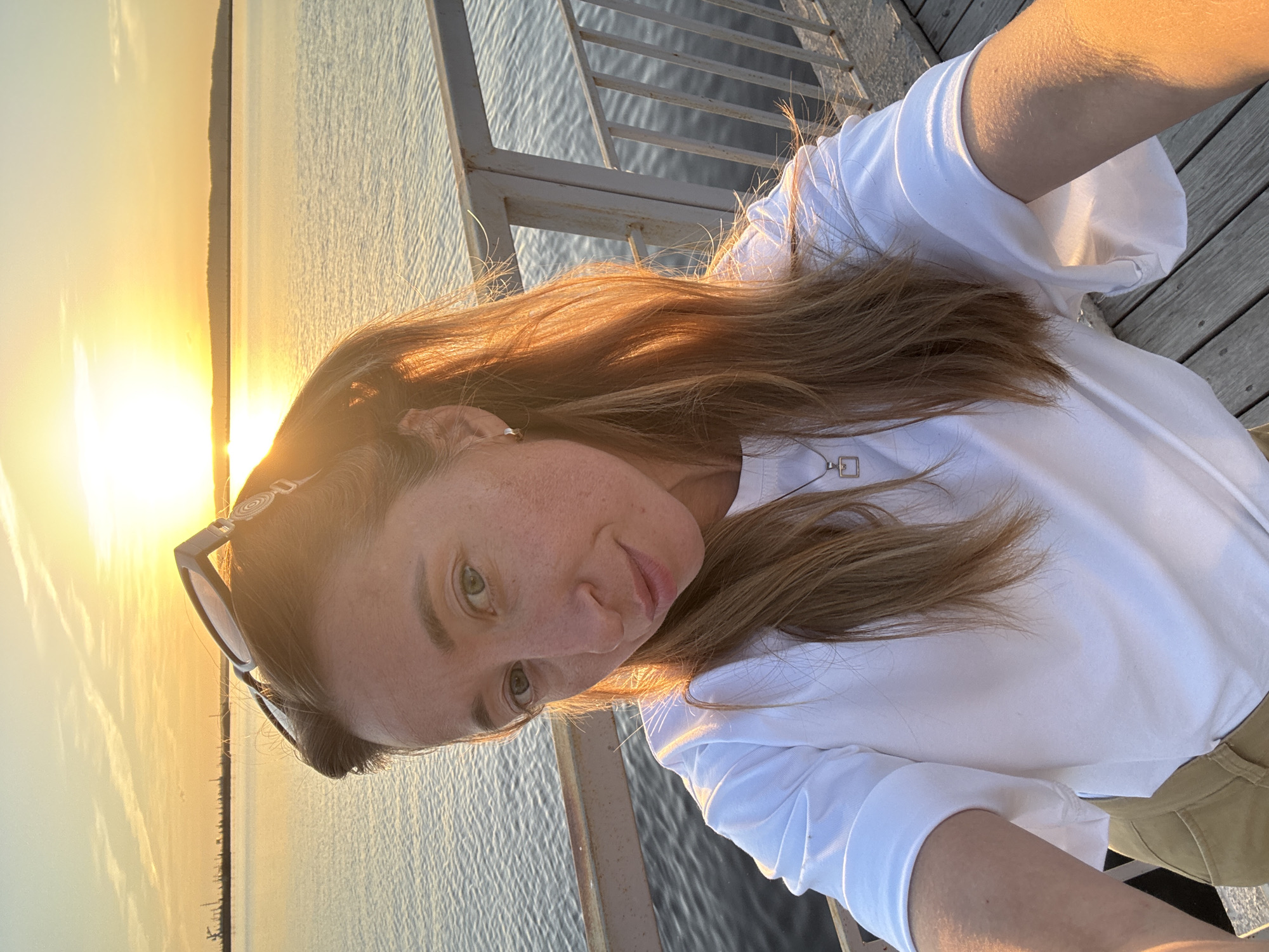 A woman with light brown hair, wearing a white long-sleeved shirt and khaki pants, stands on a pier with a metal railing. The sun is setting over a body of water. Sunglasses are perched on her head. The location is Chaikovsky, Russia.